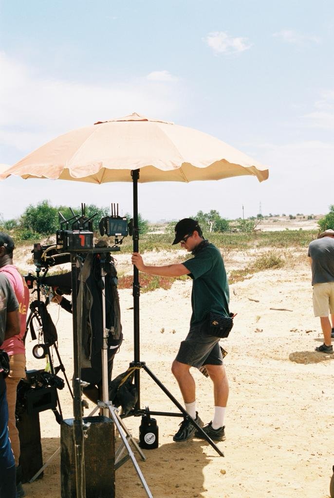 A crew member setting up equipment under a large beige umbrella on a sandy desert landscape with a few trees and sparse vegetation.