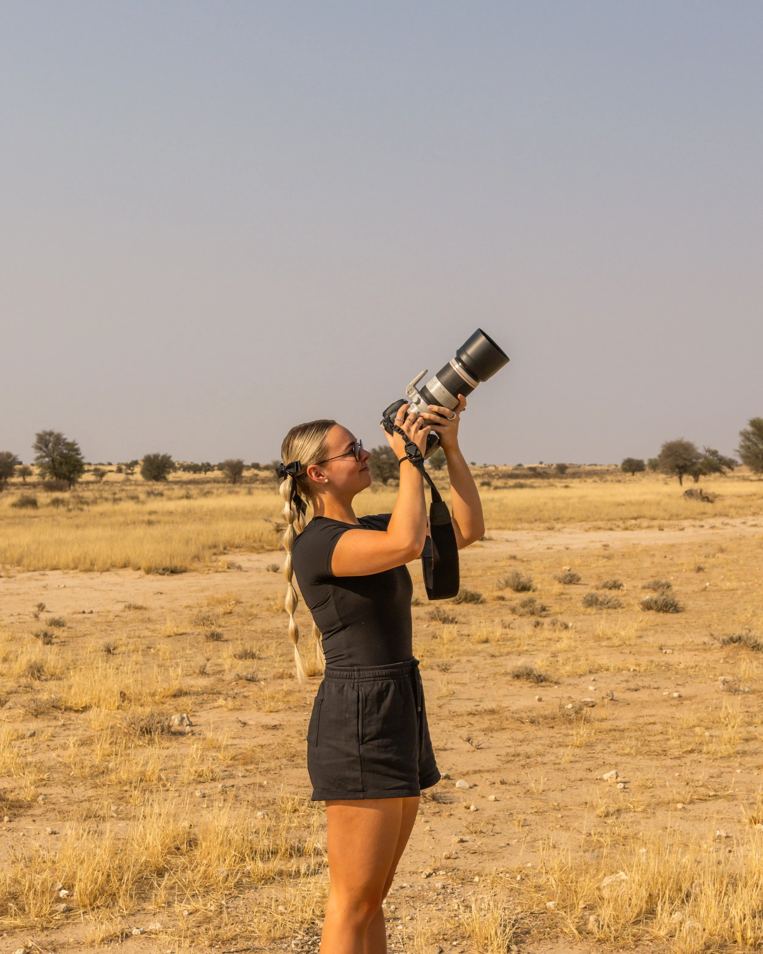 A woman with blonde hair in a braid and glasses stands in a dry, grassy field during daytime, holding a camera with a large telephoto lens pointed upward. There are a few trees and bushes in the background under a clear sky.