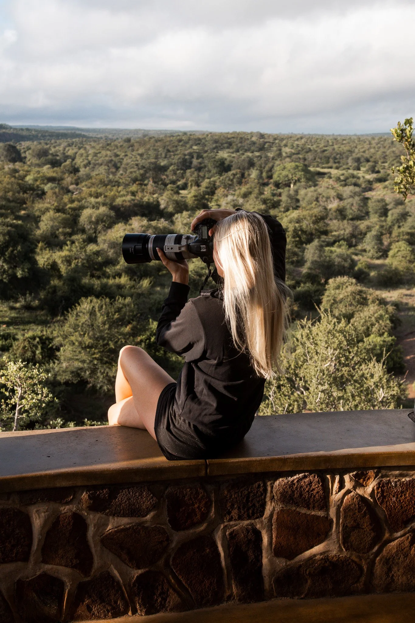 A woman with long blonde hair sitting on a stone ledge, looking through a camera telescope at a scenic landscape of trees and hills under a cloudy sky.