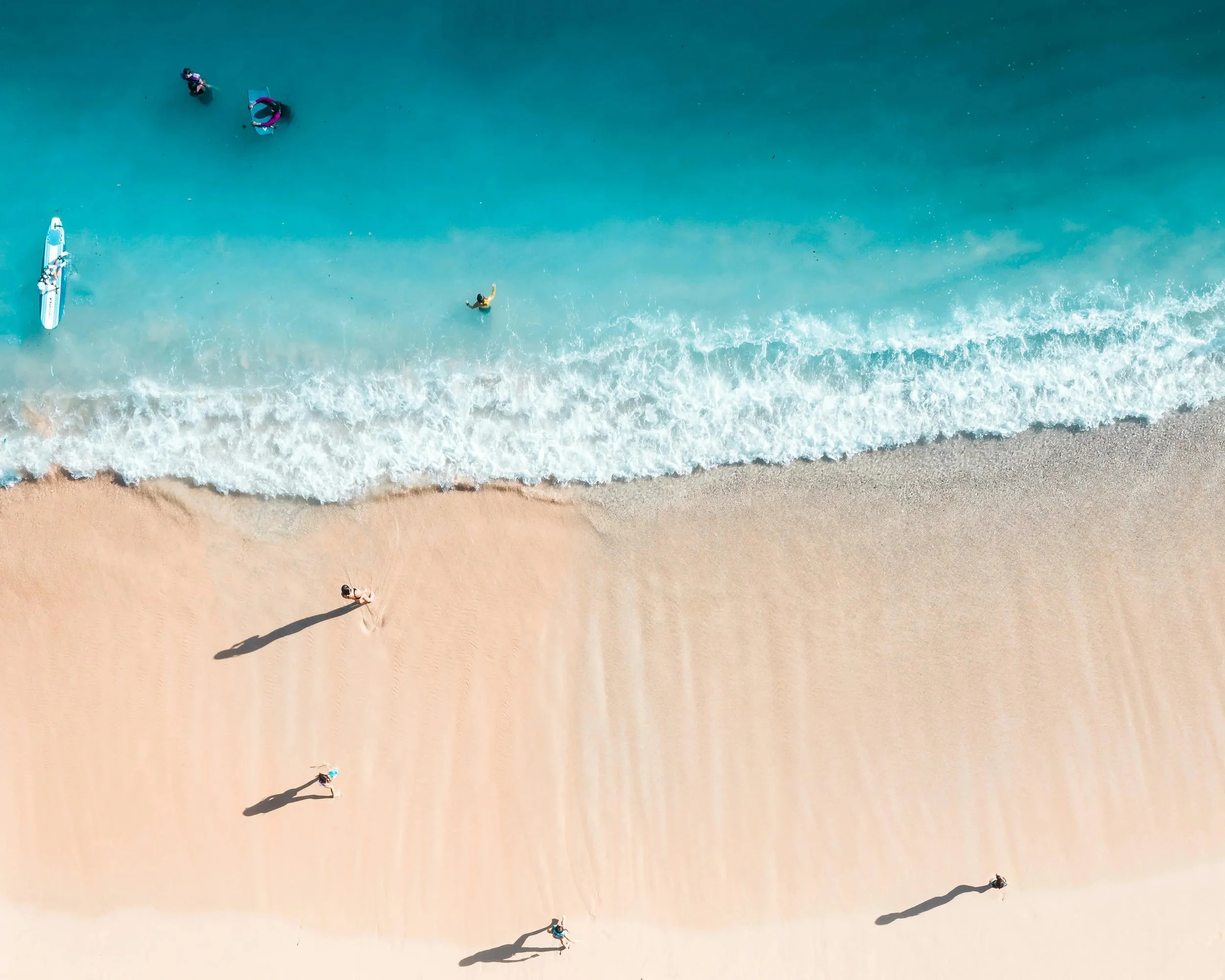 An aerial view of four people walking along the sandy beach near the water's edge, with deep blue ocean waves and turquoise water, and a paddleboard lying on the sand.