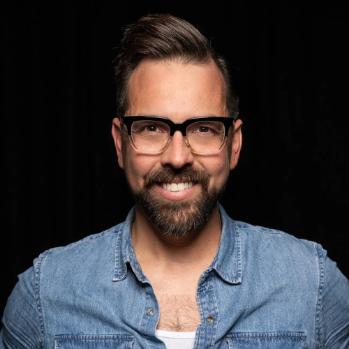 Man smiling wearing glasses and a denim shirt against a black background.