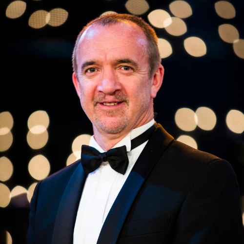 A man in a tuxedo with a bow tie, smiling, in front of a background with blurred lights.