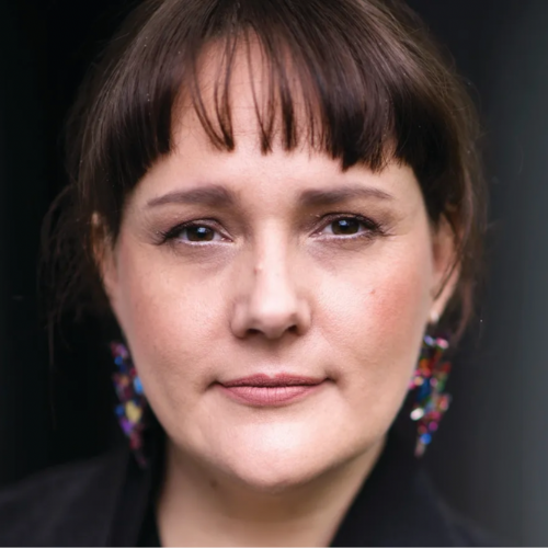 Close-up portrait of a woman with short brown hair and colorful earrings, looking directly at the camera.