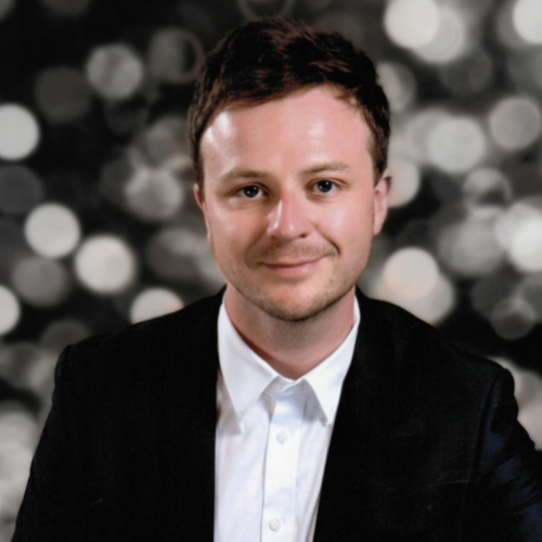 Portrait of a young man with short brown hair, wearing a black suit and white shirt, smiling against a blurred background with circular lights.