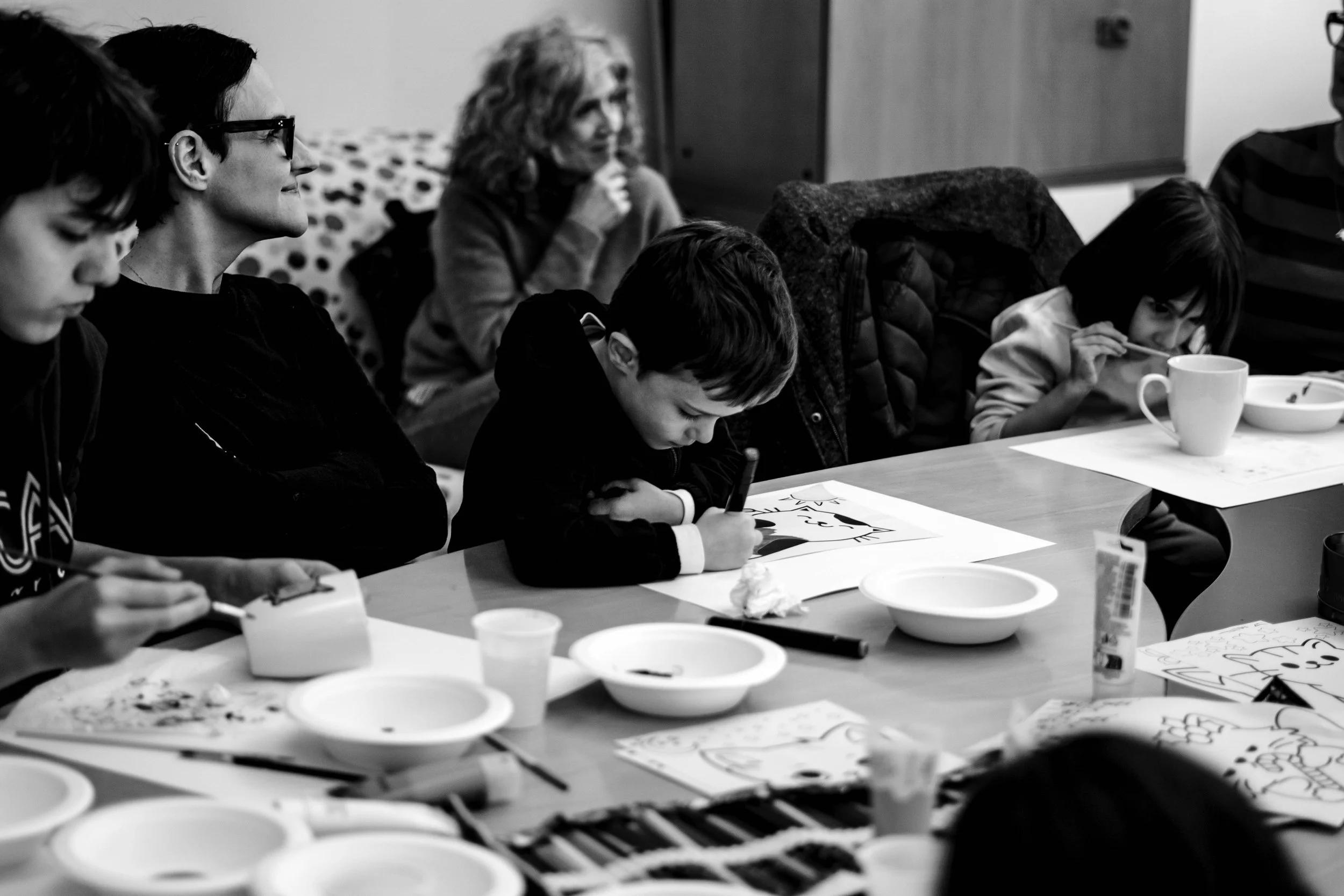 Bambini e adulti seduti a un tavolo, alcuni disegnano e altri mangiano, in un ambiente interno.