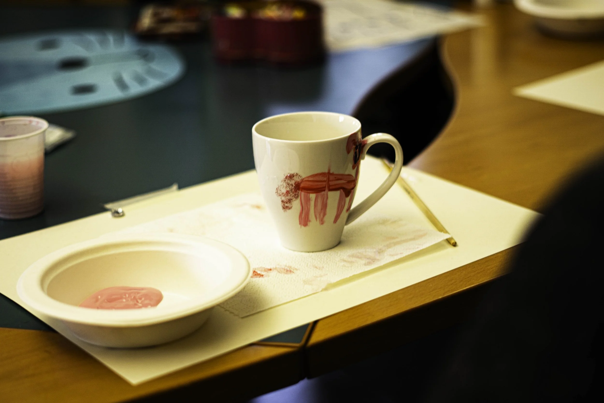 Tazza bianca decorata con disegni rossi di un animale, posizionata su un foglio di carta sotto una tovaglietta. Sul tavolo ci sono anche una ciotola di plastica con residui di pittura rosa e altri oggetti artistici.