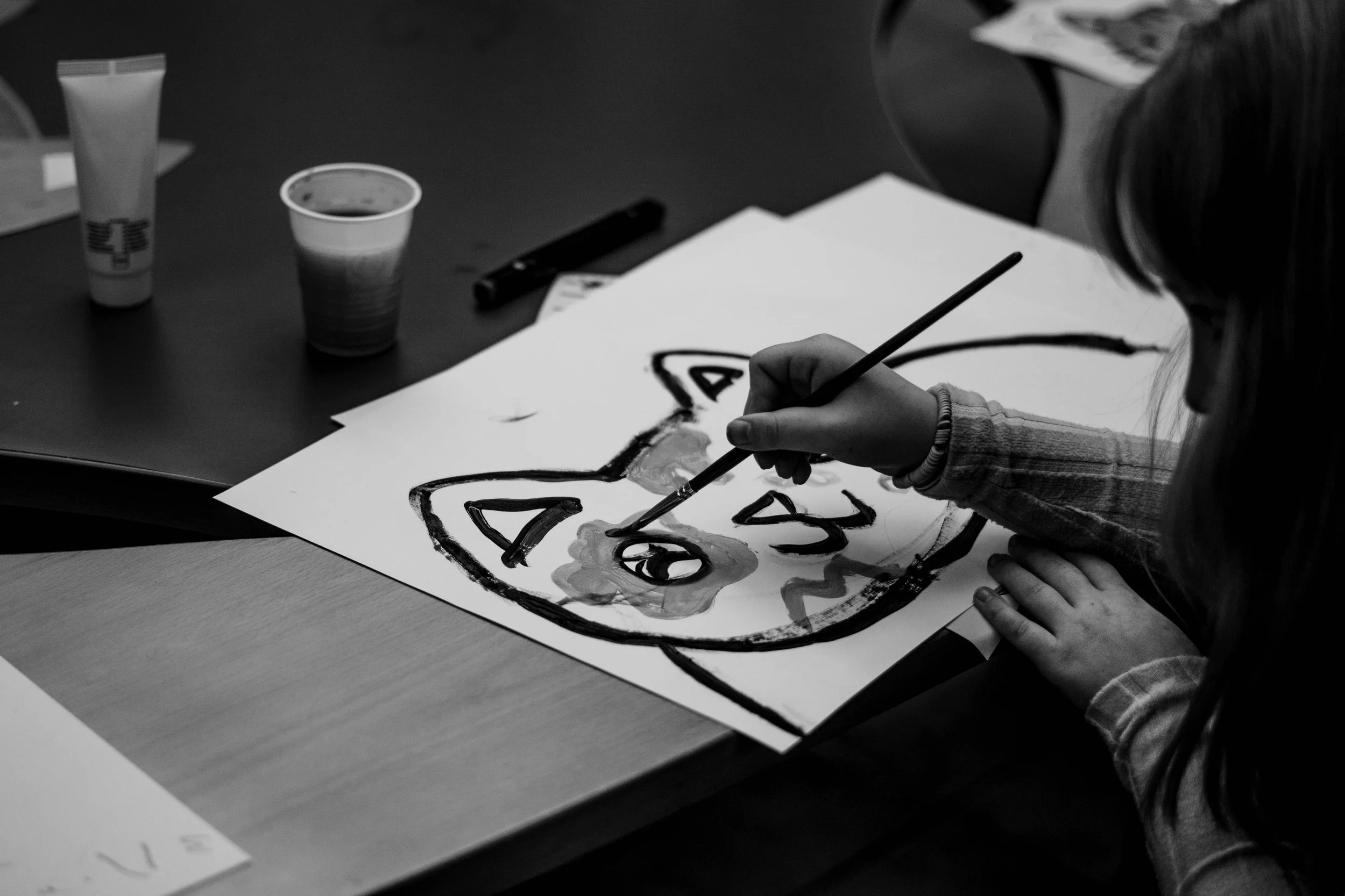 Una bambina che dipinge un disegno di un gatto con colori e pennello su un foglio di carta su un tavolo.