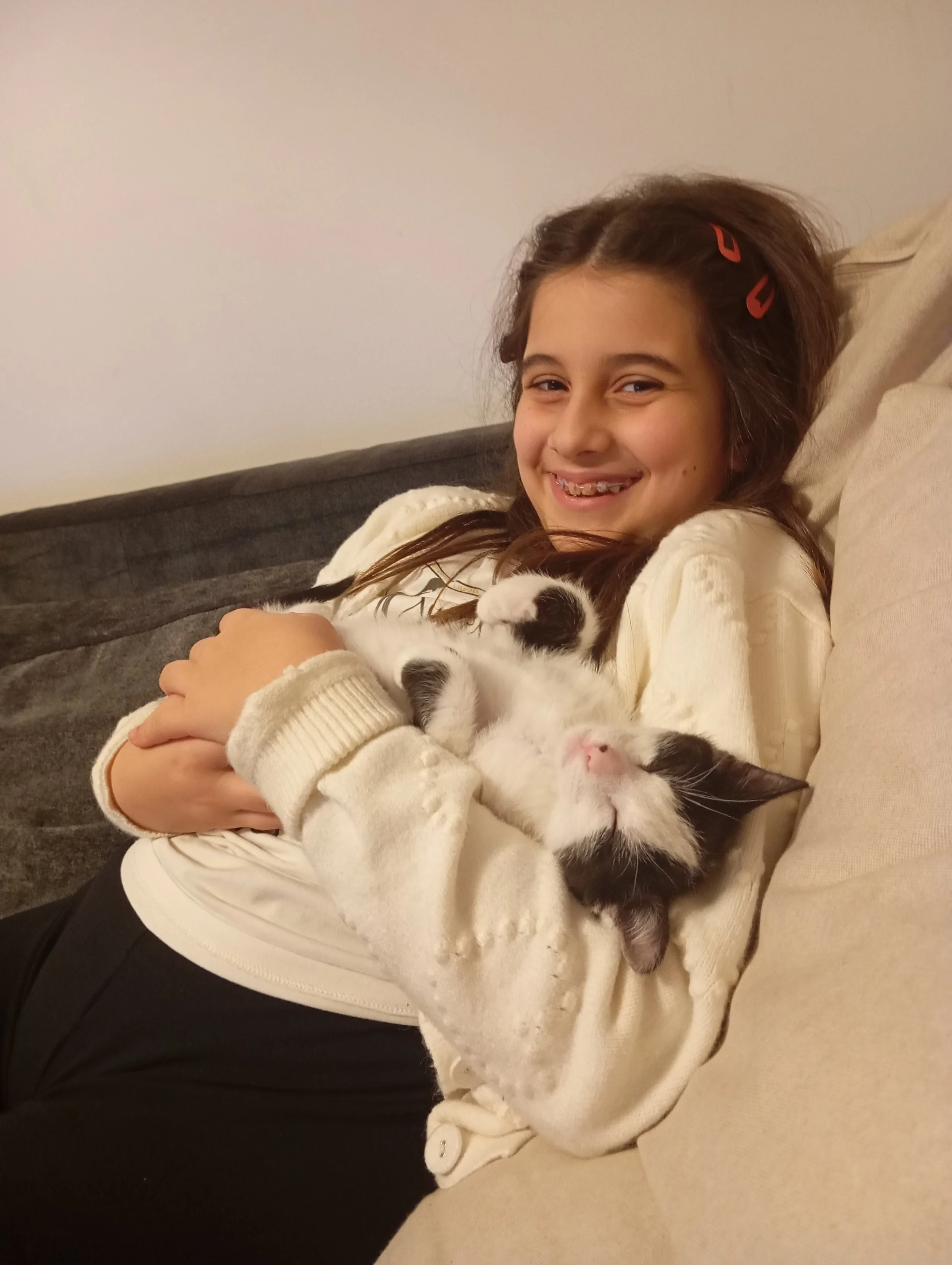Una ragazza sorridente di circa 10 anni, con capelli castani e due mollette rosse, abbraccia un gatto bianco con macchie nere, che giace sul suo petto, entrambi seduti su un divano in un ambiente domestico.