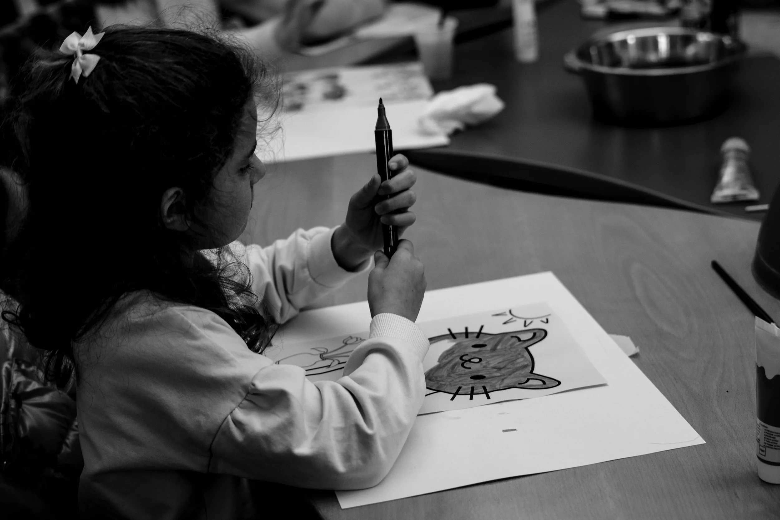 Una bambina seduta a un tavolo che sta colorando un disegno di un orso con un sole, in bianco e nero.