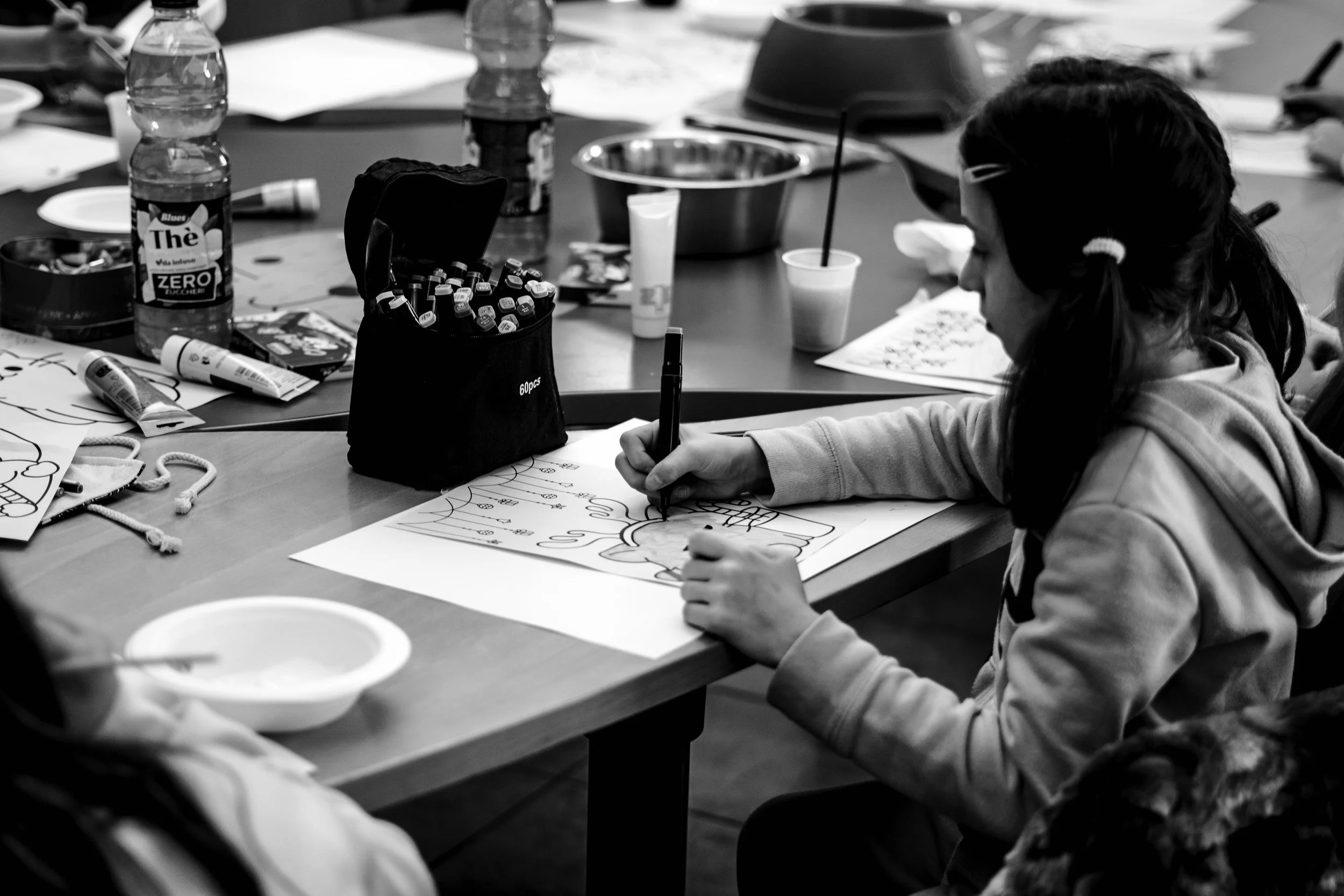 Una bambina che disegna un'immagine colorata su un foglio, seduta a un tavolo con vari oggetti come bottiglie, colori e fogli sparsi.