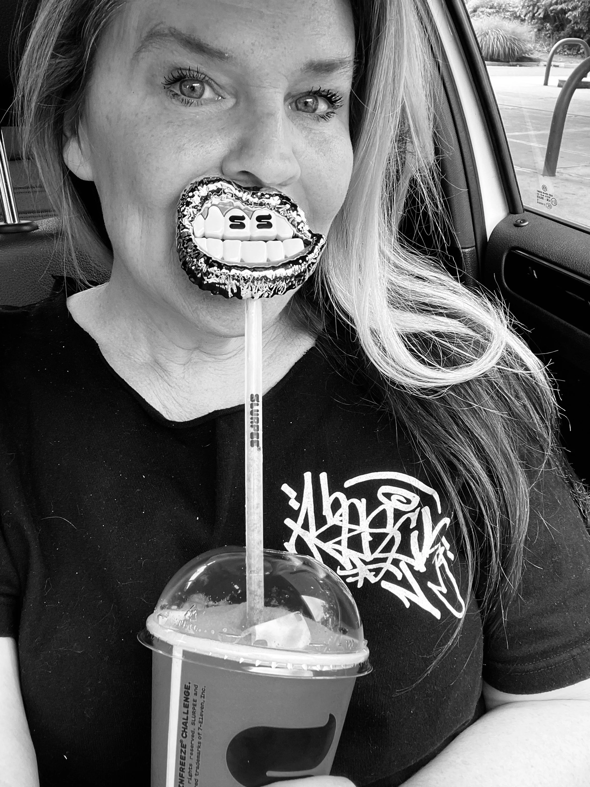 A woman with long hair and a black shirt holding a large lollipop with a cartoon mouth design, sitting inside a car in a parking lot.