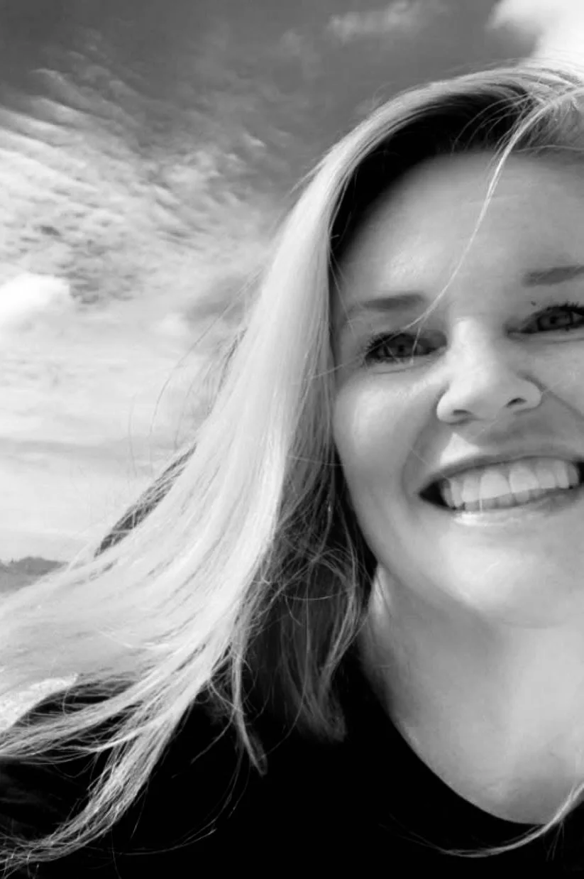 A woman with long hair and a black shirt holding a large lollipop with a cartoon mouth design, sitting inside a car in a parking lot.