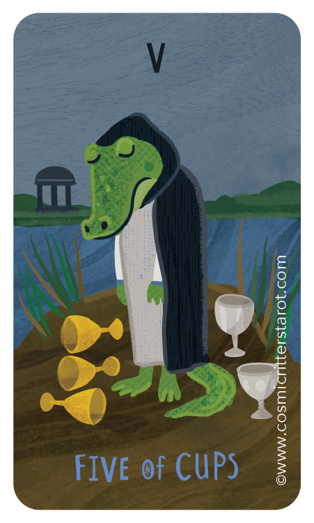 A cartoon alligator with a human body wearing a white robe and black cape, standing by water with tall grass, five gold cups on the ground, and a small building and trees in the background. The card reads 'FIVE OF CUPS' and has a 'V' at the top.