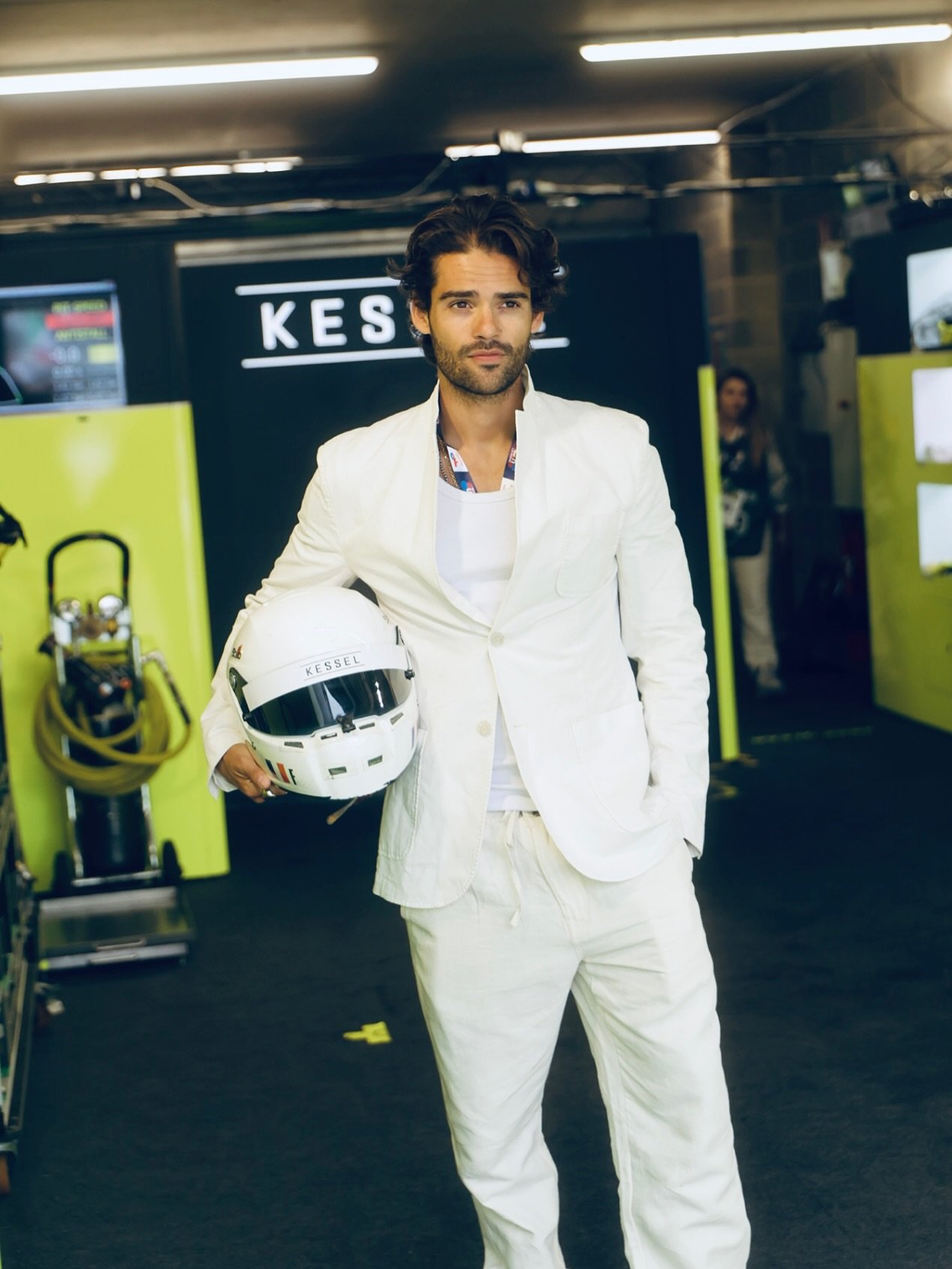 A man in a white suit holding a white motorcycle helmet, standing in a parking garage with a sign that reads 'KESS' in the background.