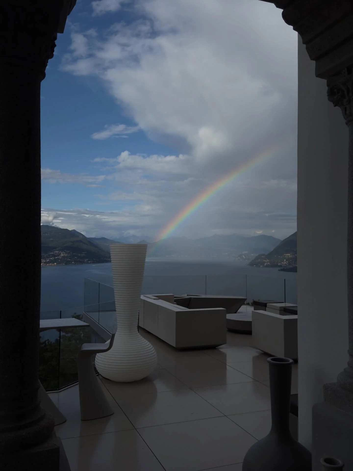 View of a patio with modern white furniture and a large vase, overlooking a lake with mountains in the background, a rainbow arching across the sky, viewed through an arched doorway.