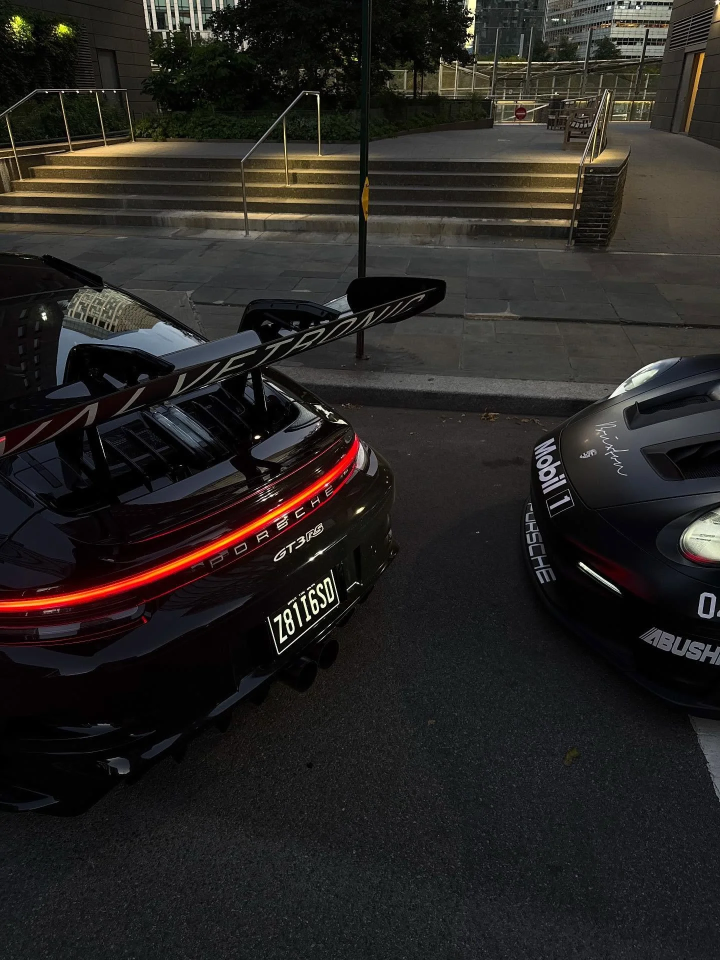 Black Porsche 911 GT3 RS with racing decals and a large rear wing, parked next to a gray Porsche 911 Carrera with racing decals. Both cars are in an urban setting near steps leading to a building.