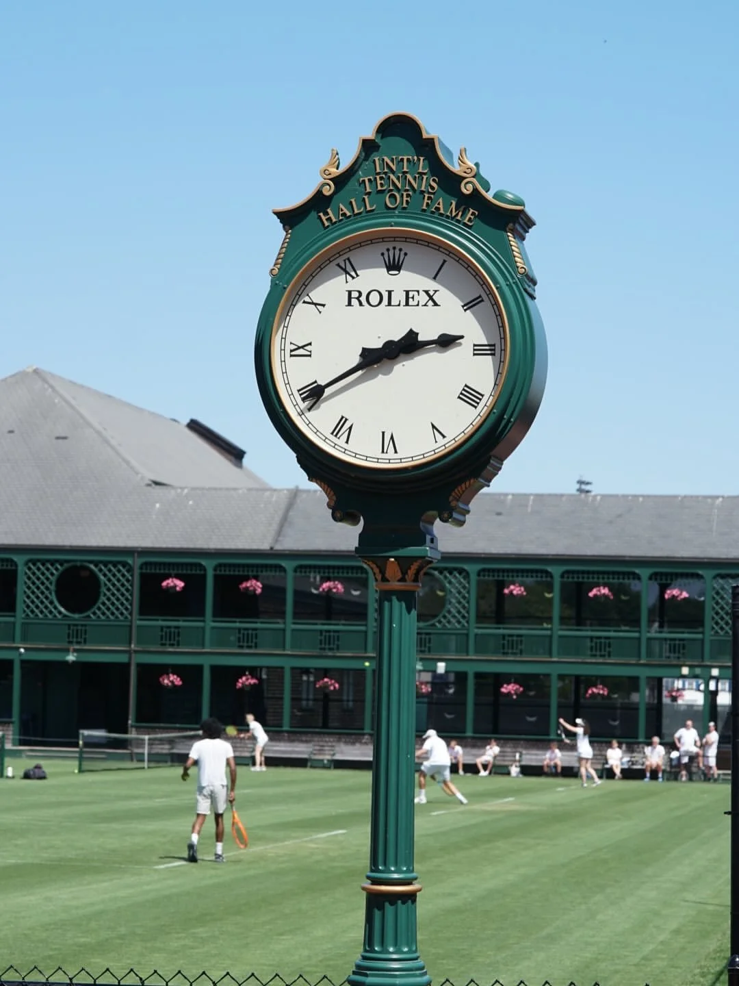 A large clock with the Rolex logo and Roman numerals, labeled 'International Tennis Hall of Fame,' overlooking a tennis court with players practicing in the background.