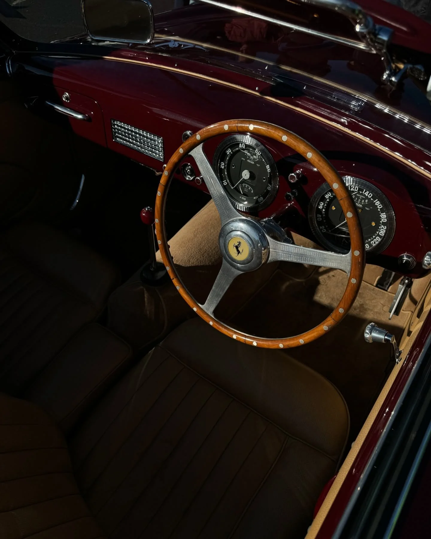 Interior of a vintage Ferrari car showing a wooden steering wheel with a Ferrari logo, dashboard with analog gauges, and brown leather seats.