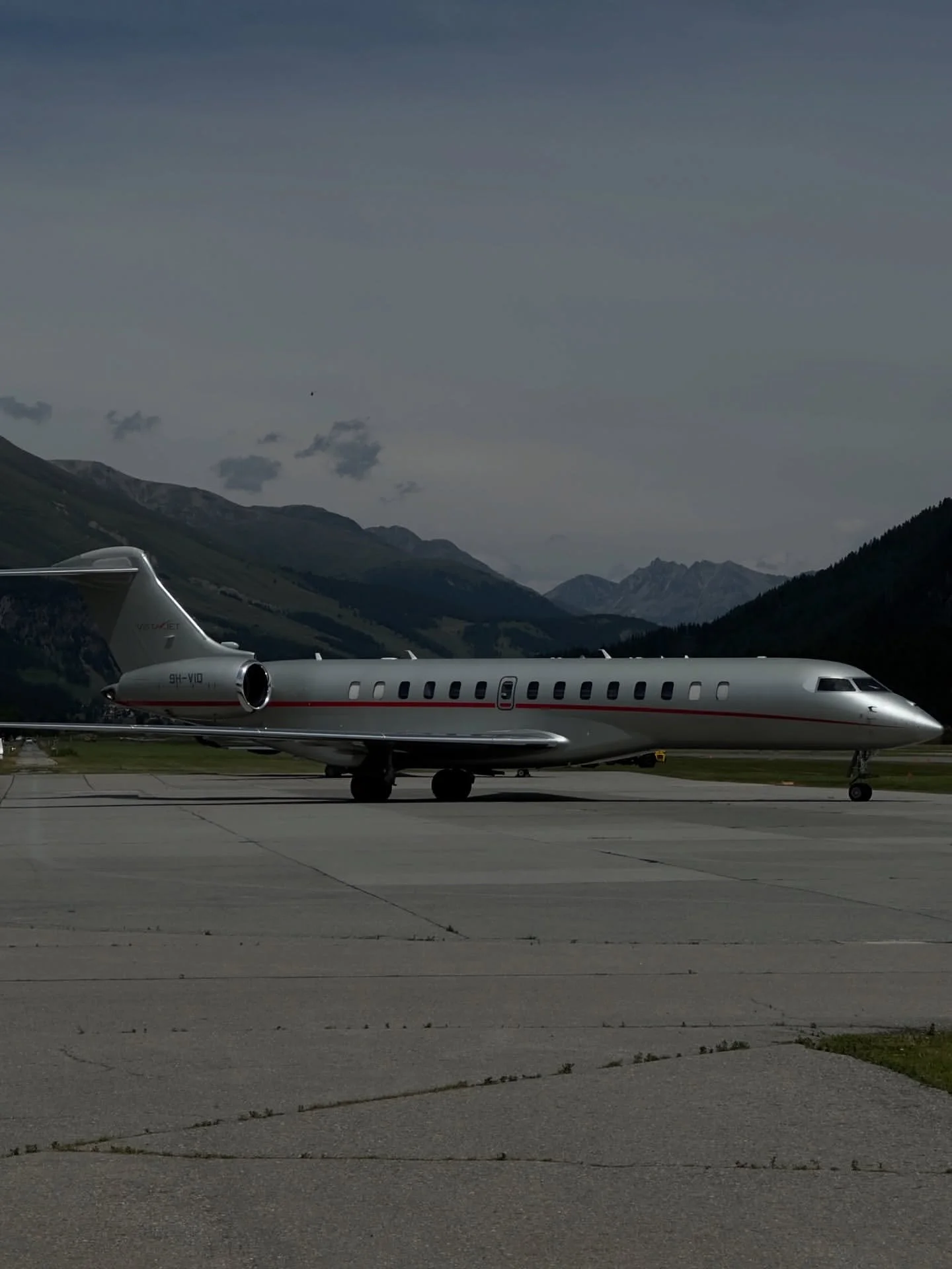 A sleek, silver private jet on an airport tarmac with mountains in the background under a cloudy sky.