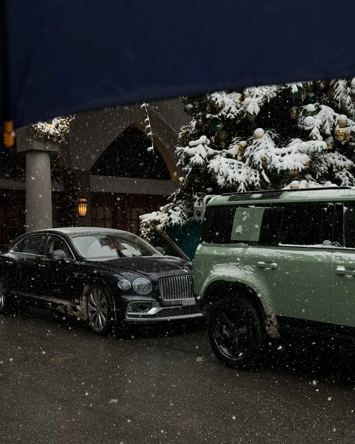 Snow falling on parked cars outside a building with a large Christmas tree decorated with gold ornaments.