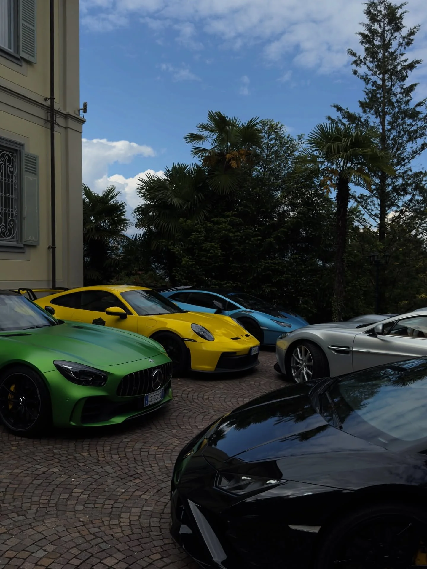 Multiple luxury sports cars parked on a cobblestone driveway with a building on the left and trees in the background under a partly cloudy sky.