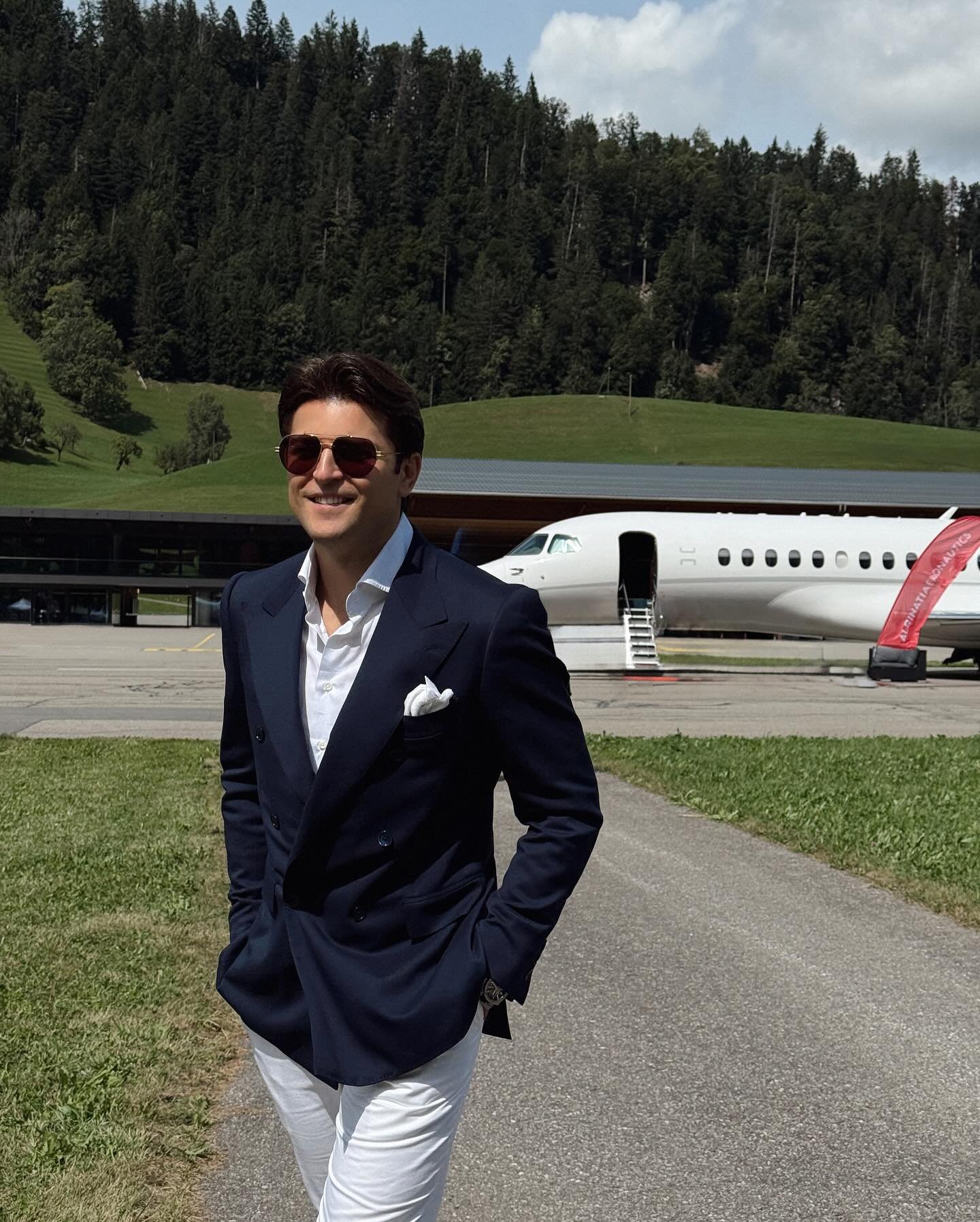 A young man in a navy blazer, white shirt, white pants, and sunglasses standing outside near a private jet aircraft, with green hills and trees in the background.