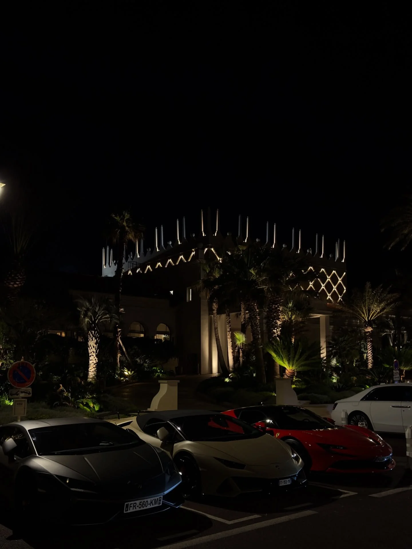 Night view of luxury cars parked in front of a brightly lit building with palm trees.