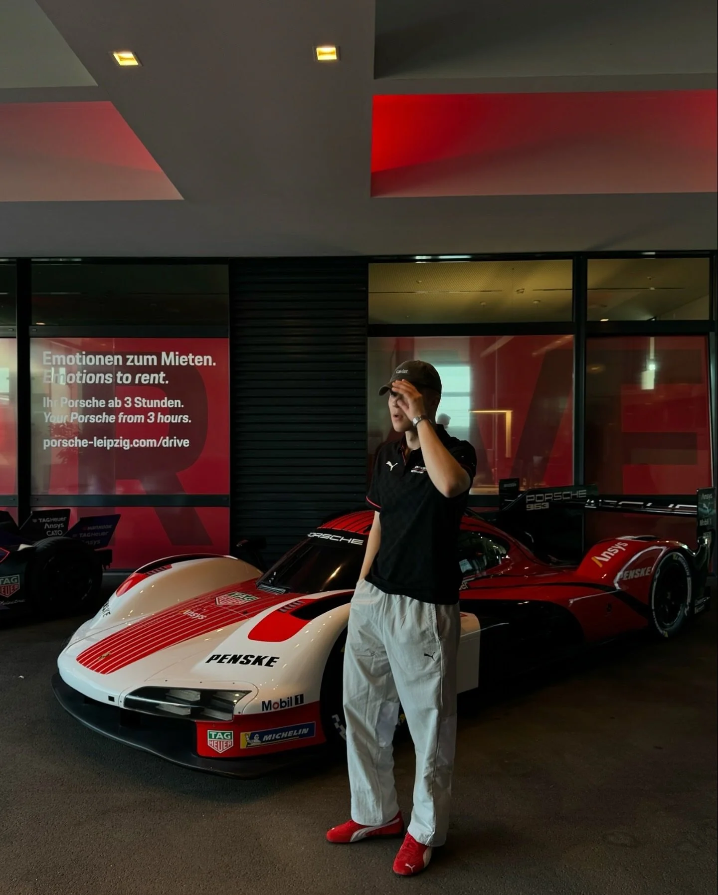 A person standing in front of a vintage racing car at a car dealership, with a sign in German and English promoting Porsche rentals.