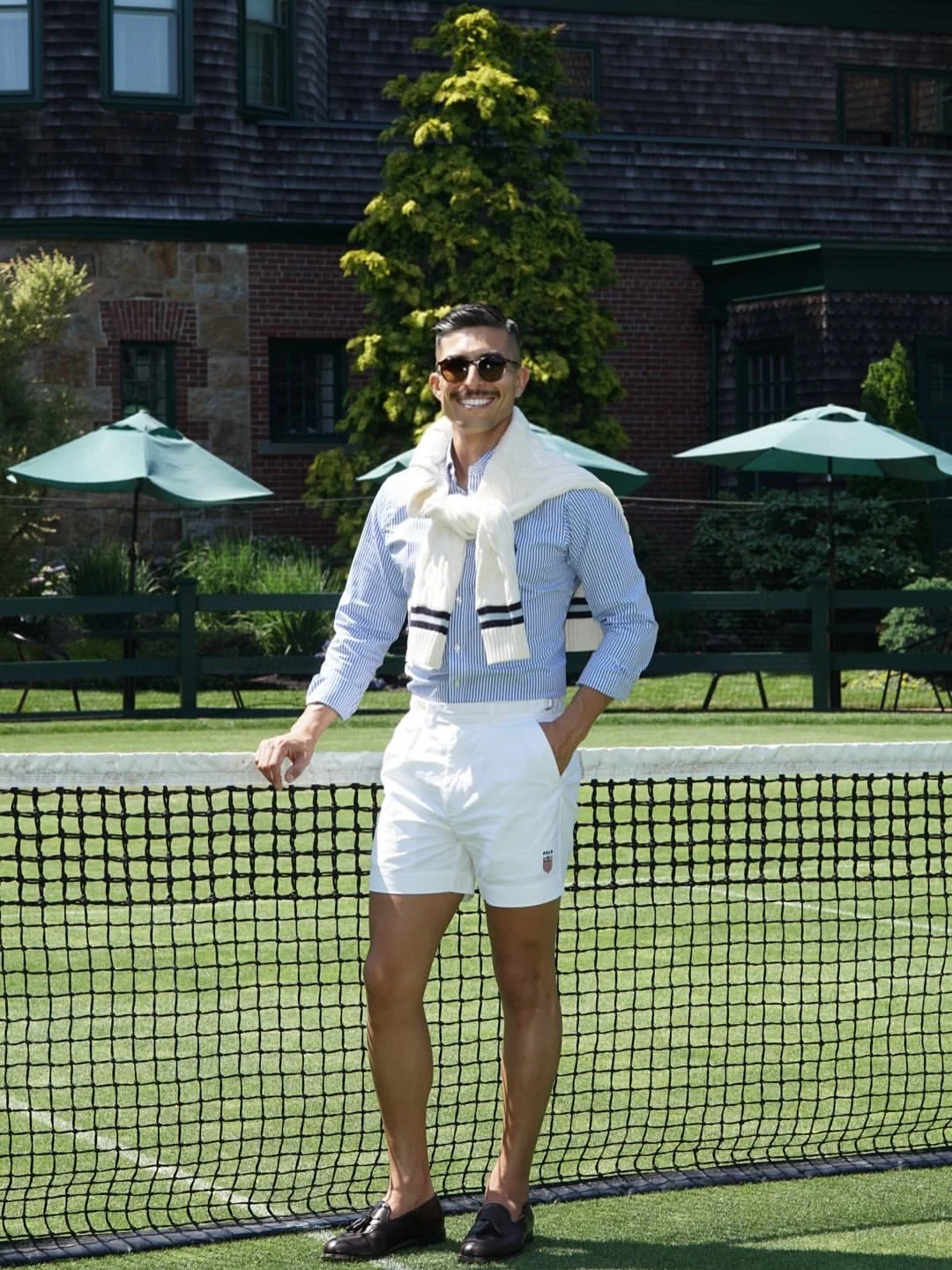 A man standing on a tennis court, smiling, wearing sunglasses, a striped shirt, white shorts, and loafers, with a sweater tied around his shoulders.