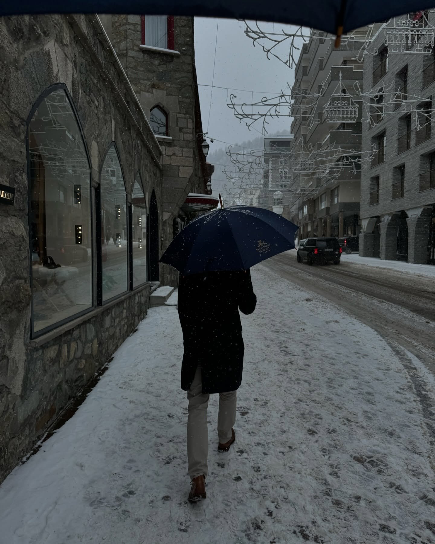 Person walking on snowy sidewalk during snowfall, carrying a blue umbrella, with buildings and street in the background.