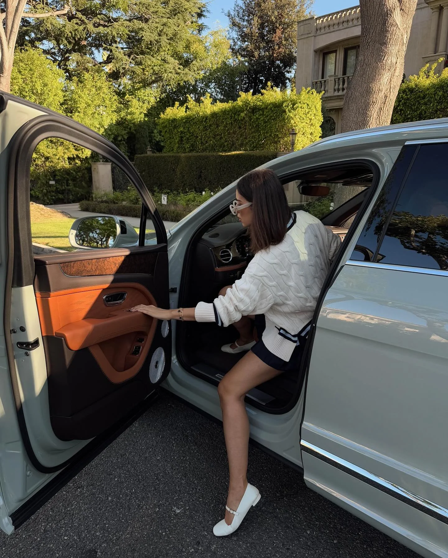 A woman in a white sweater, navy shorts, and white heels getting into a white luxury car with a brown interior, parked outdoors on a sunny day near trees and a building.