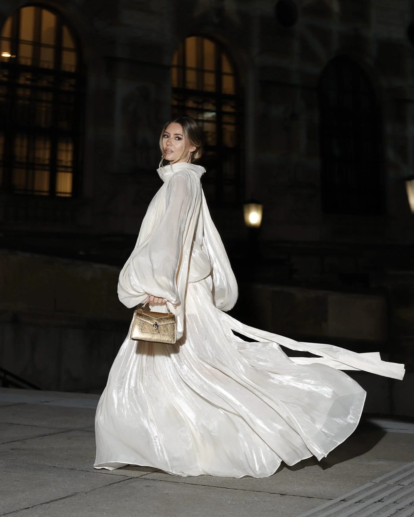 A woman dressed in a flowing white gown carrying a gold handbag, standing on city sidewalk at night with historic building and streetlights in background.