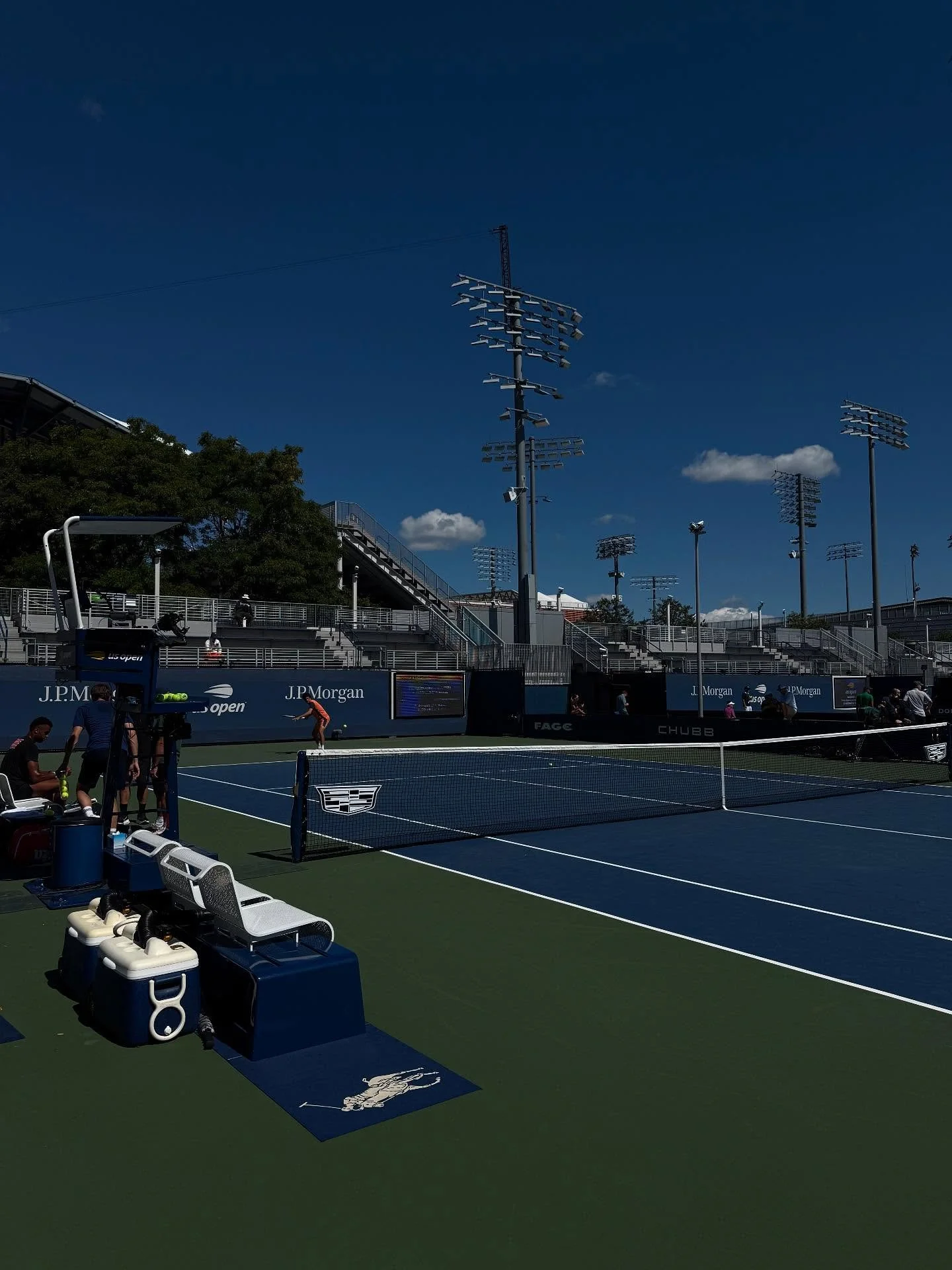 A tennis court with players practicing, surrounded by tall floodlights and seating stands under a clear blue sky.