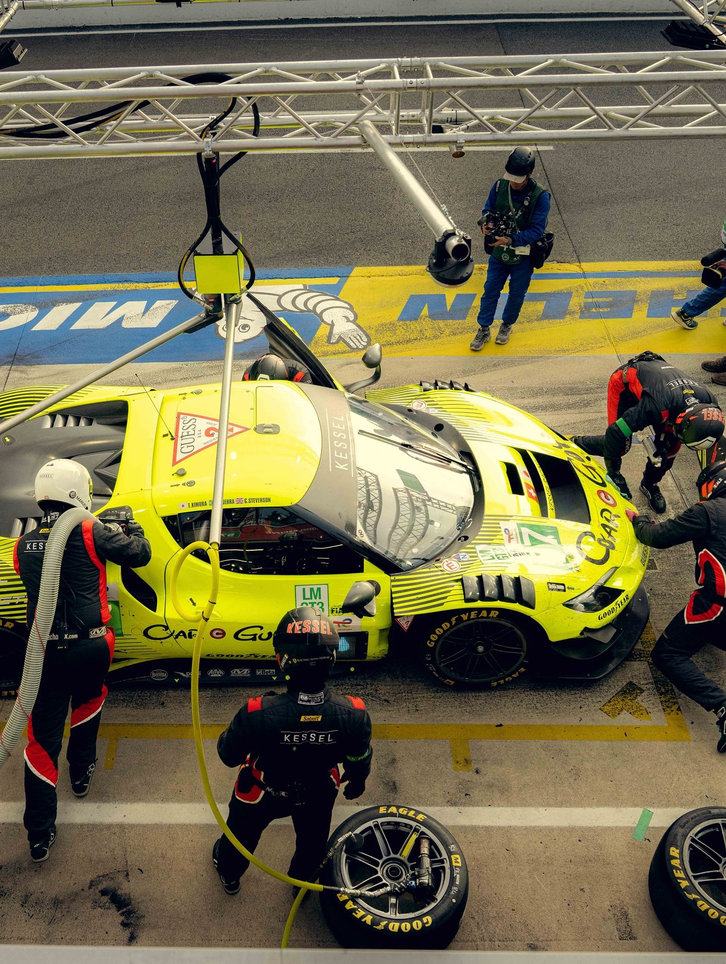 Race car pit stop with a yellow racing car in the pit lane surrounded by crew members changing tires and working on the vehicle.