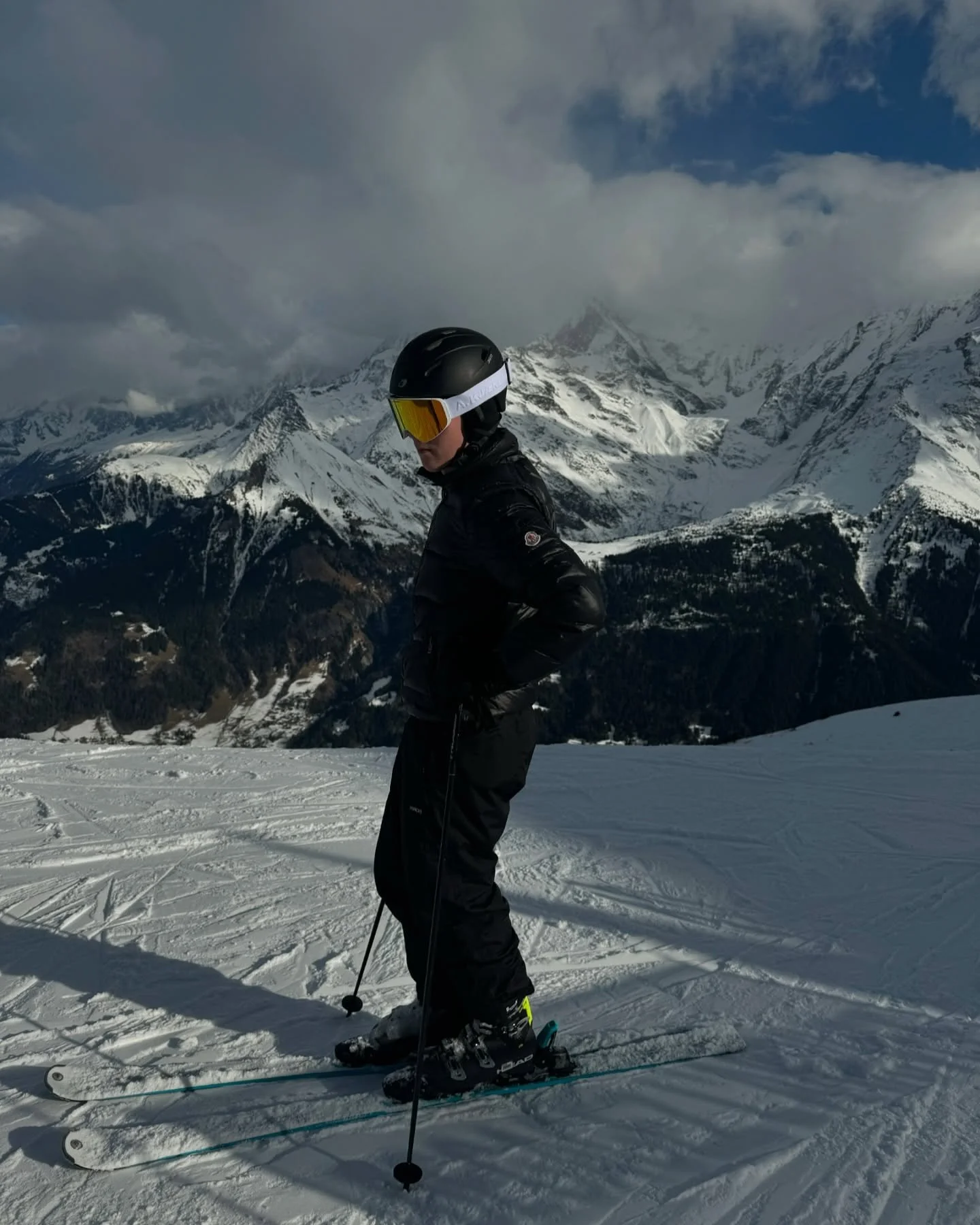 A person wearing ski gear, including a helmet and goggles, standing on snow-covered slopes with snow-capped mountains in the background.