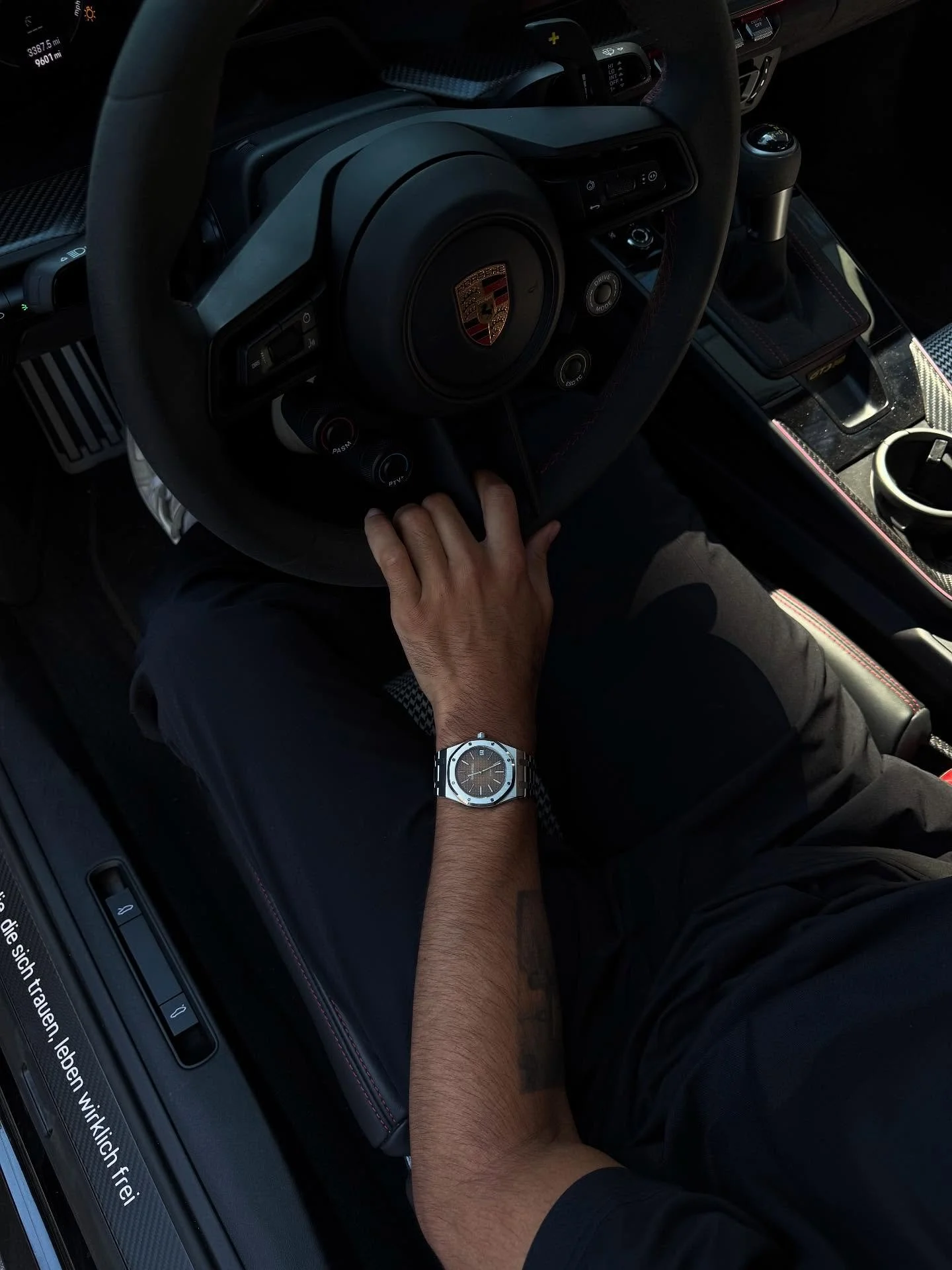 A person sitting in the driver's seat of a Porsche sports car, with their left hand on the steering wheel and wearing a silver wristwatch.