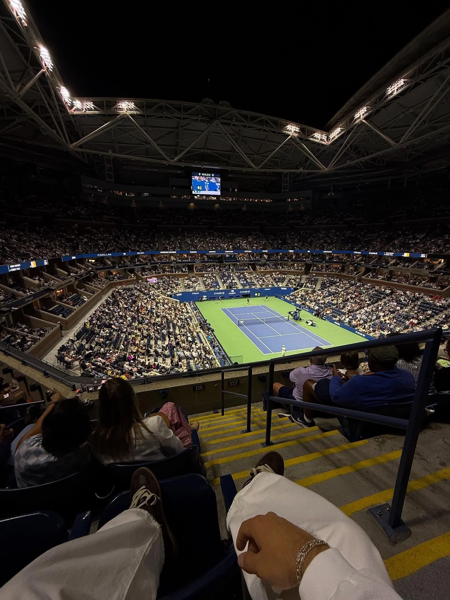 Indoor tennis stadium filled with spectators watching a match, with a view from the seats behind the court, showing the court and audience.