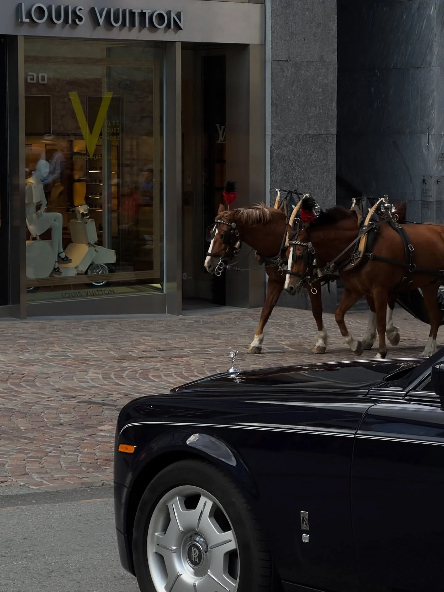 Two brown horses with harnesses pulling a black luxury car on cobblestone street in front of Louis Vuitton store.