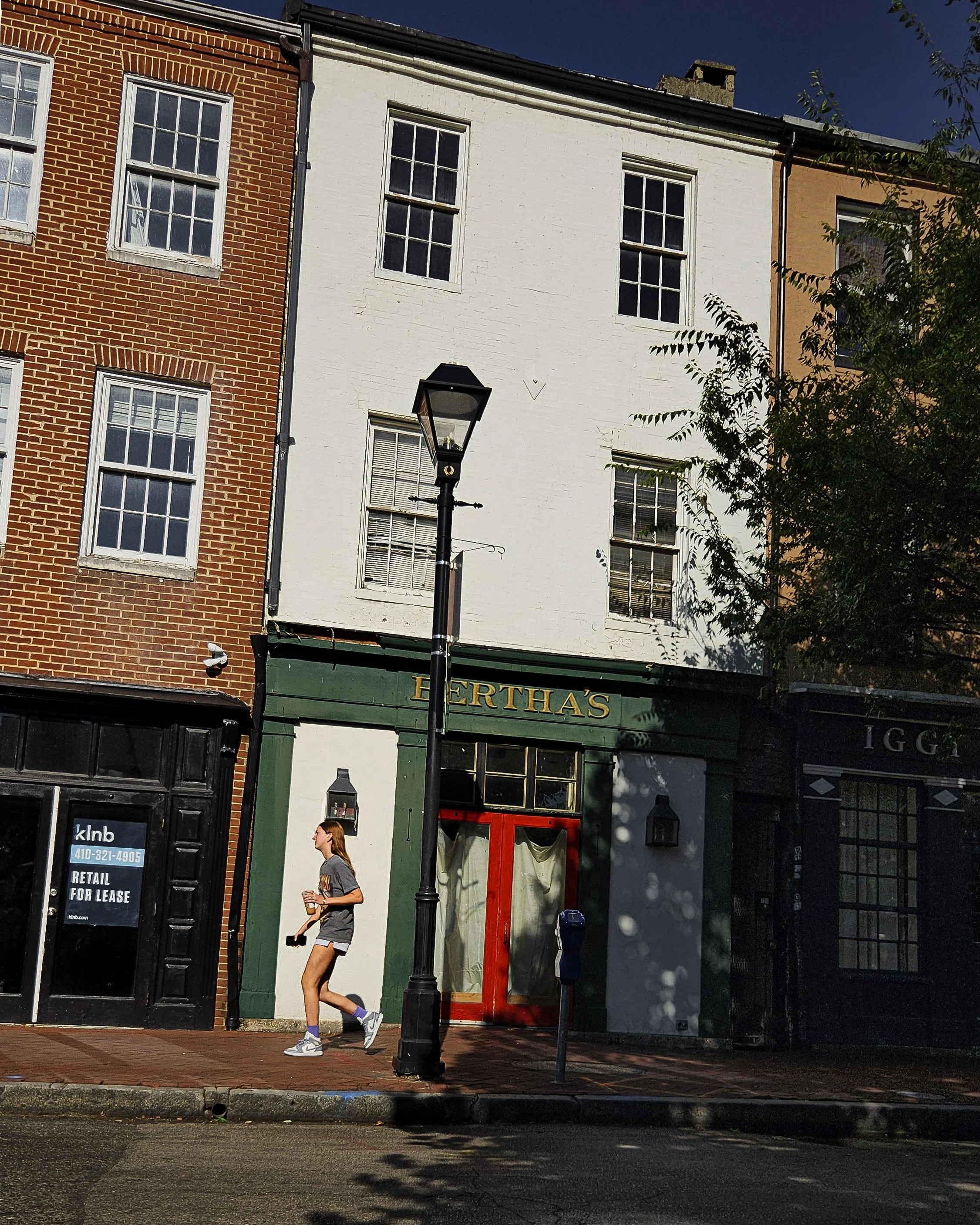 A woman walks past a storefront with a green facade and red door on a city street, with brick and painted buildings, a lamppost, and trees.