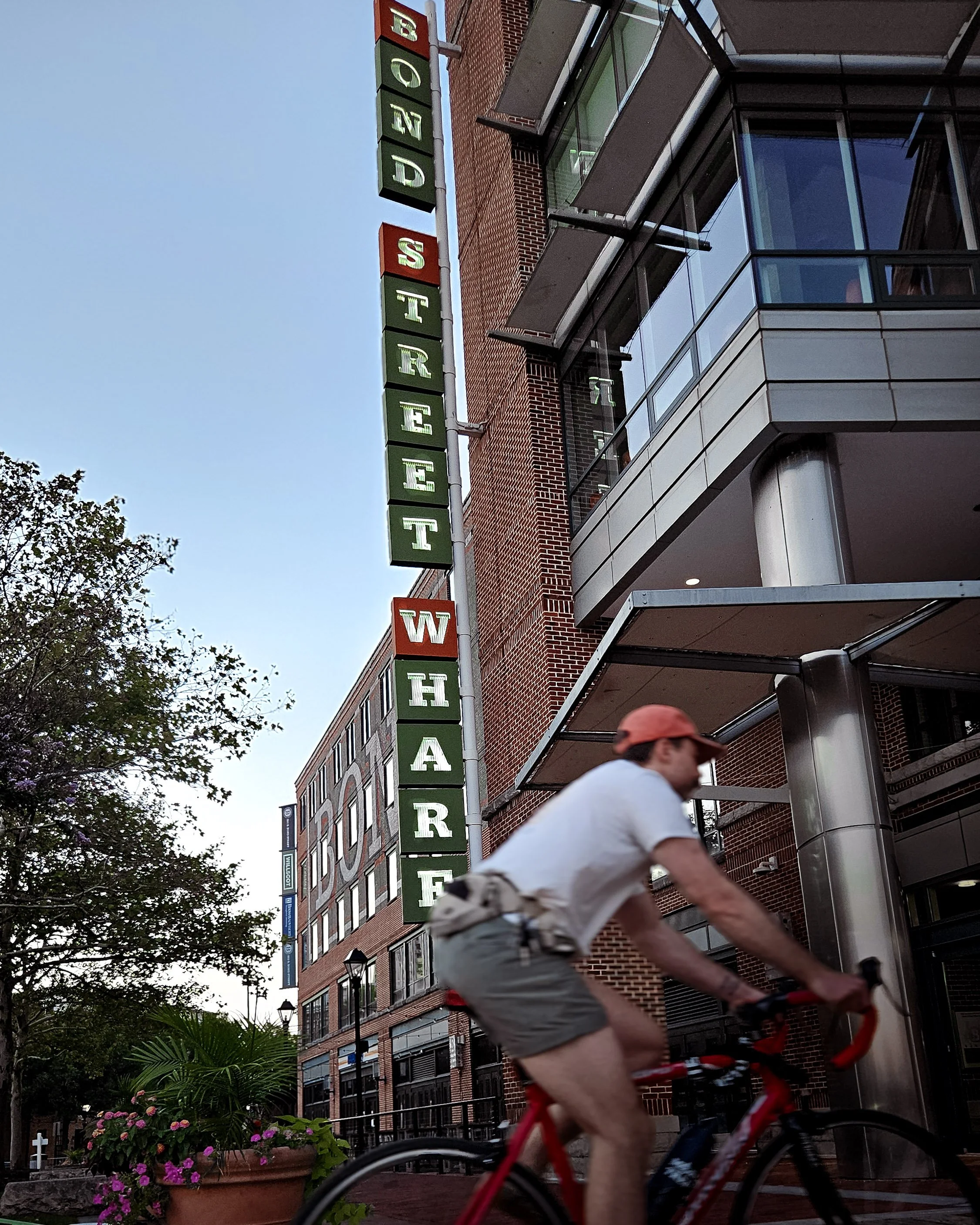A man riding a bicycle past a vertical sign for Board Street WAREHOUSE, with modern brick and glass buildings and a tree with flowers nearby.
