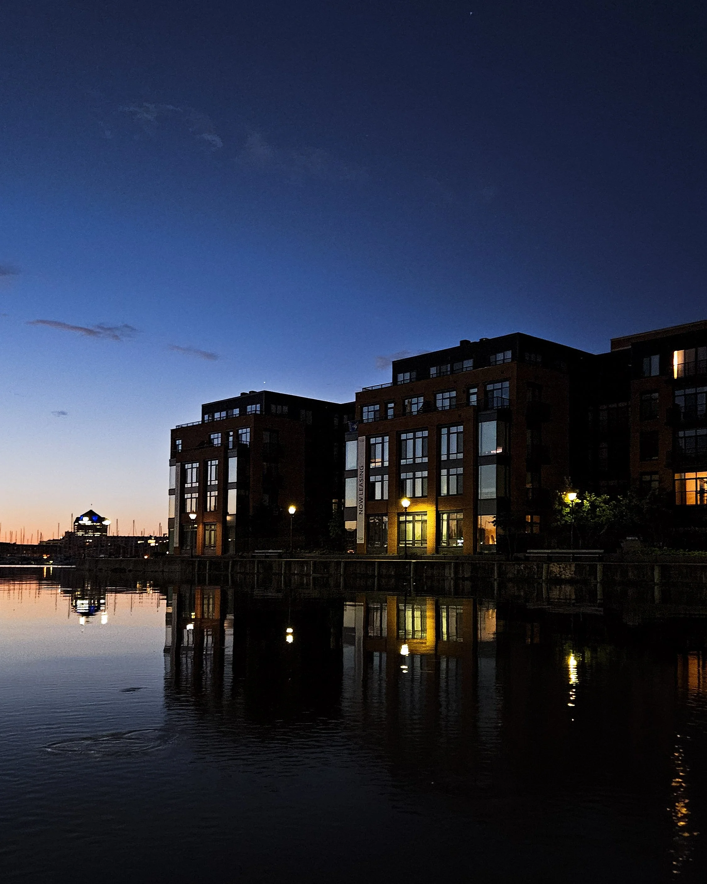 Nighttime view of modern apartment buildings along a waterfront, with reflections in the water and a clear, darkening sky.