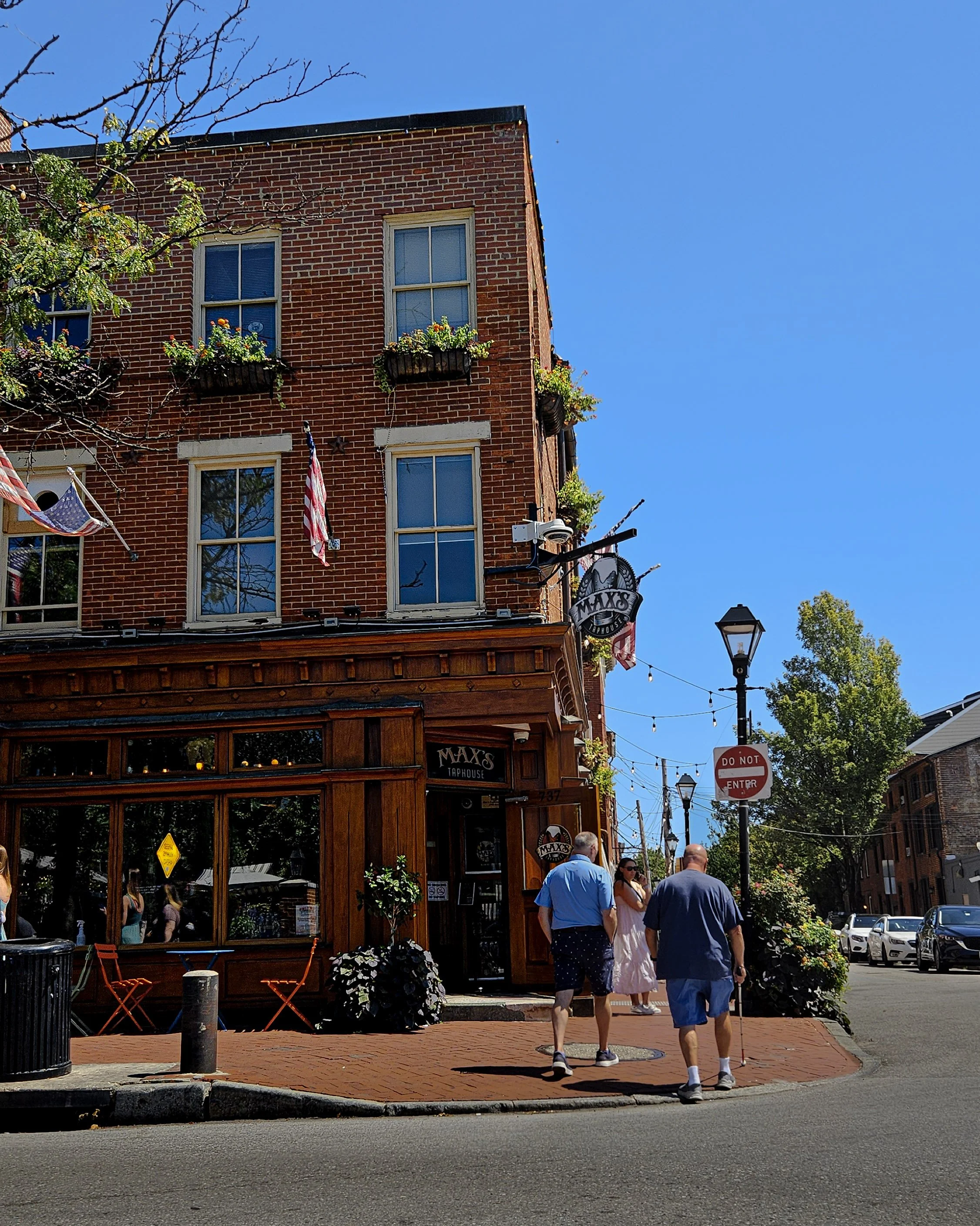 Street scene showing a brick building with a wooden storefront labeled 'Max's Thapouse', American flags, people walking on the sidewalk, and a street with parked cars under a clear blue sky.