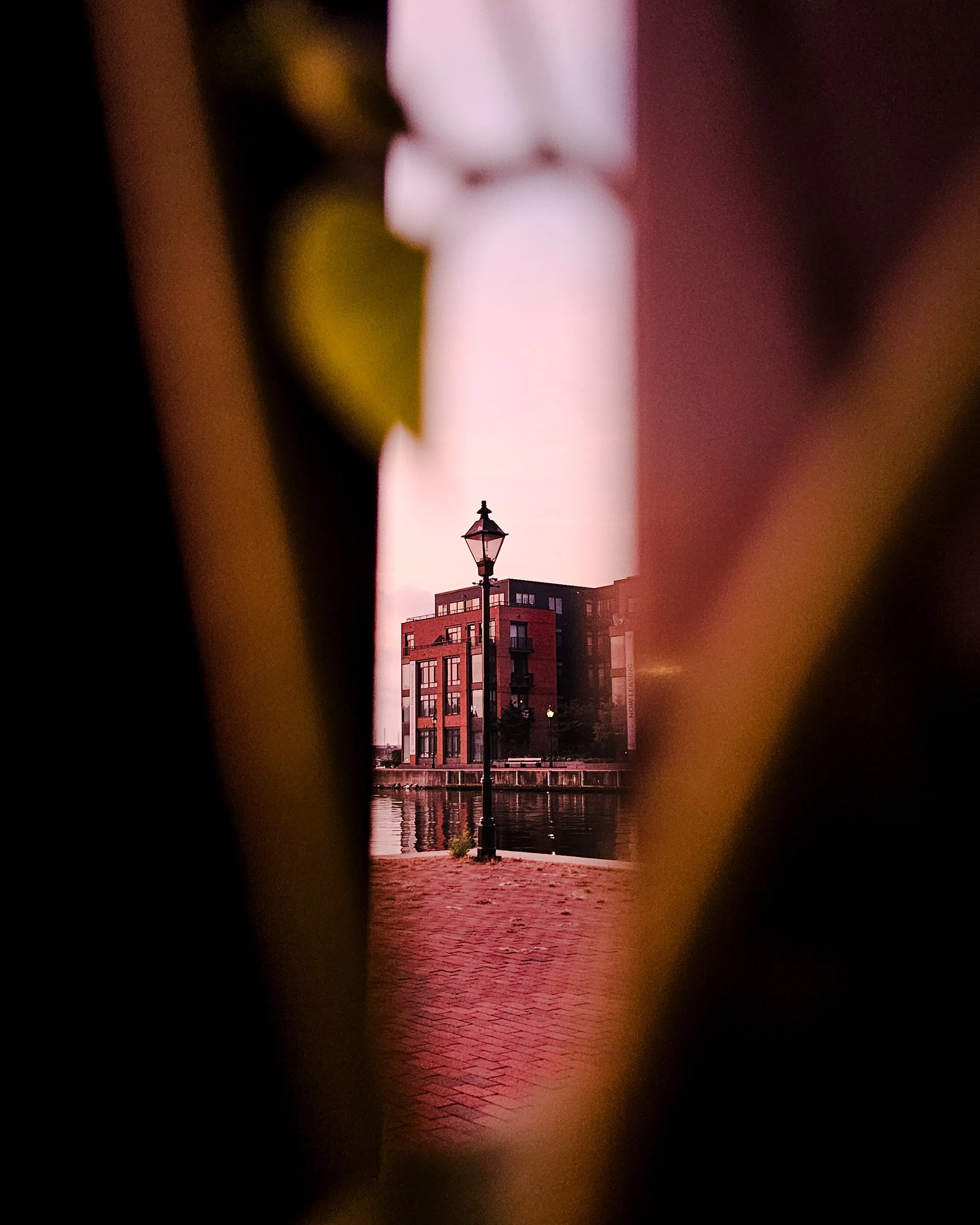 View of a building and a streetlamp through a narrowed gap, possibly between curtains or blinds, with a waterfront and urban backdrop at dusk or dawn.