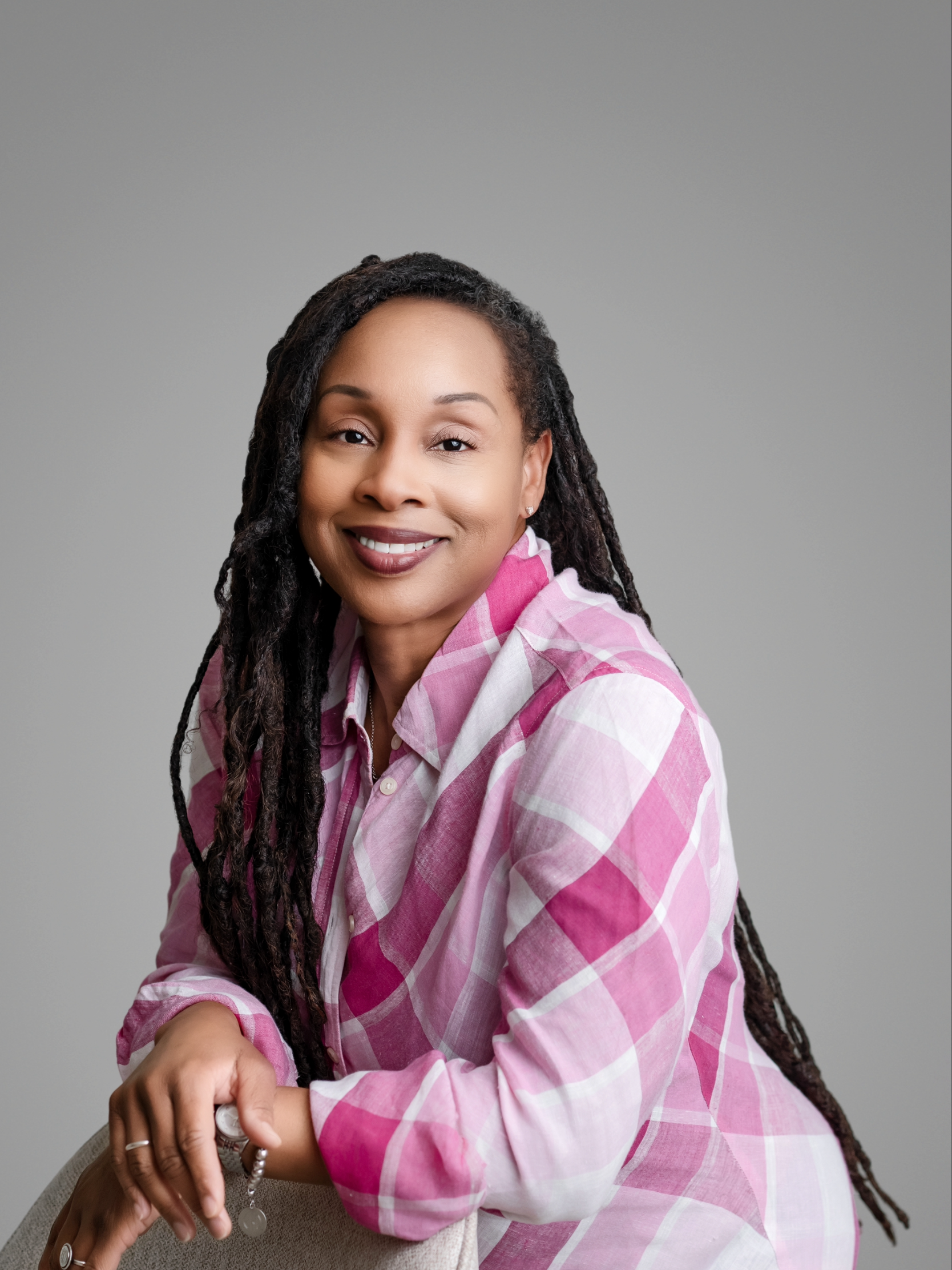 Woman with long locs wearing a pink and white plaid shirt, smiling while seated against a plain gray background.