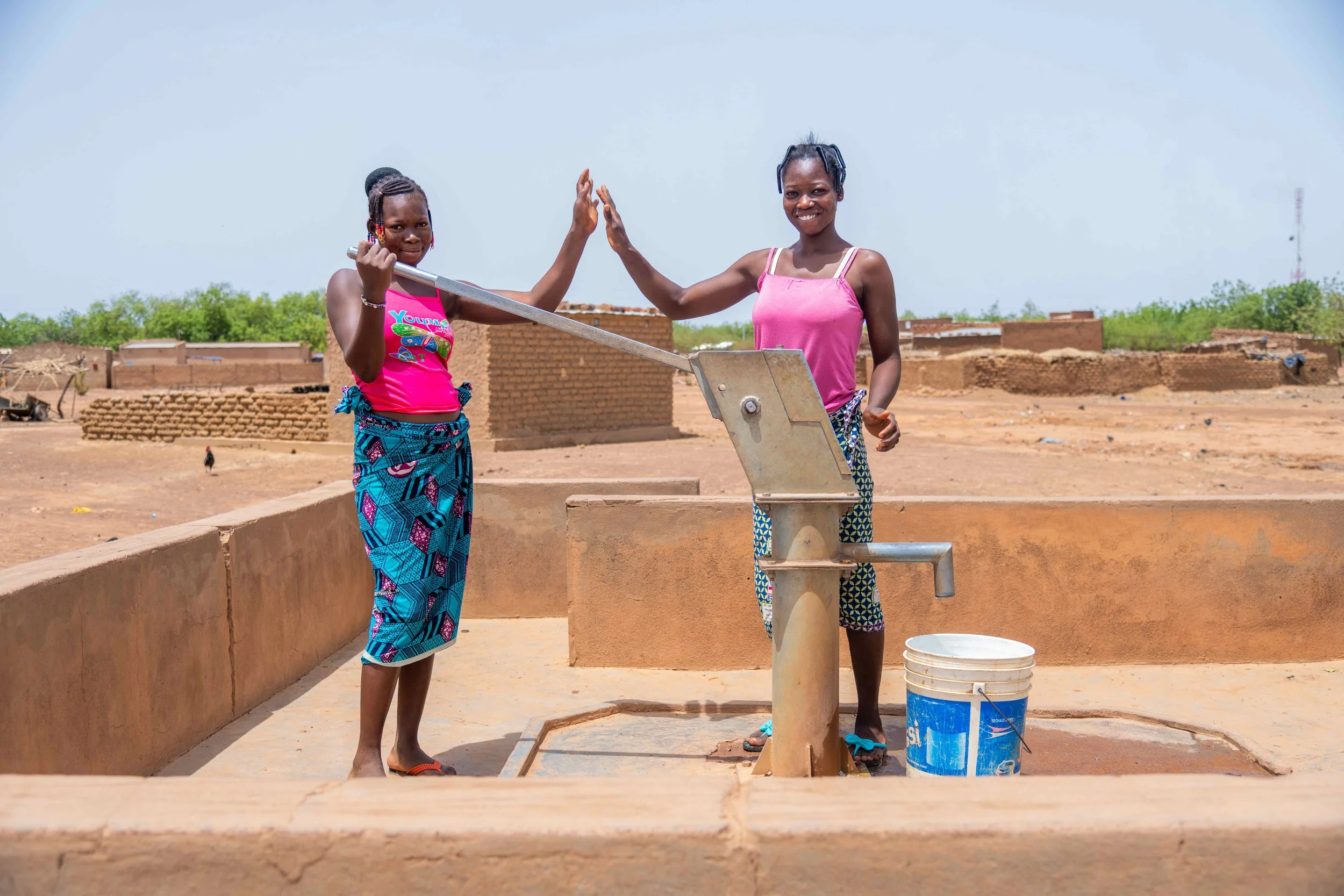 Two girls are giving each other a high five while pumping water from a well