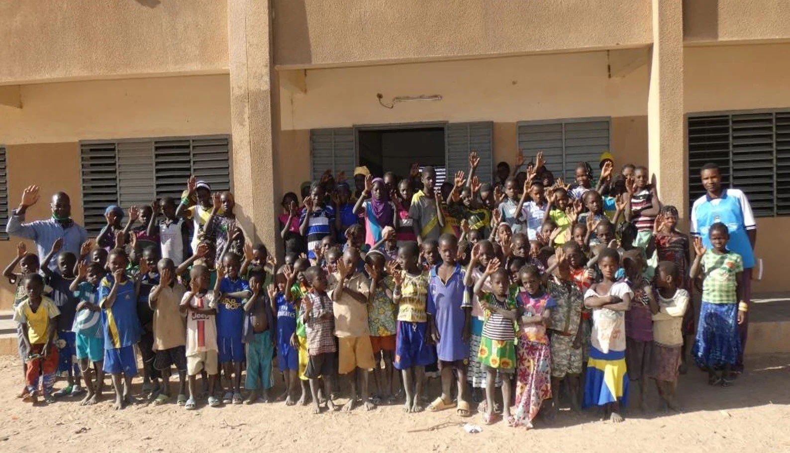 Students and teachers stand in front of their school.Some are waving at the camera