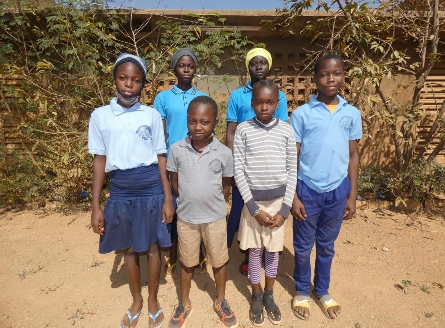 Six children are standing in front of a fence looking into the camera.
