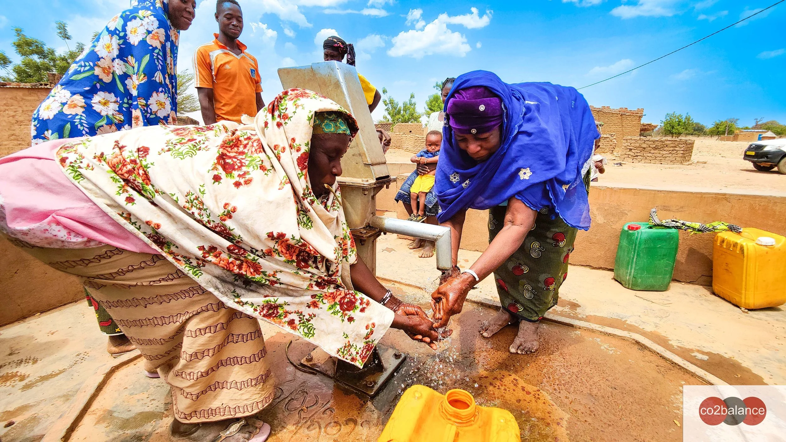 Two women are pumping water from a well. They are wearing brightly coloured clothes. The sky is bright blue.