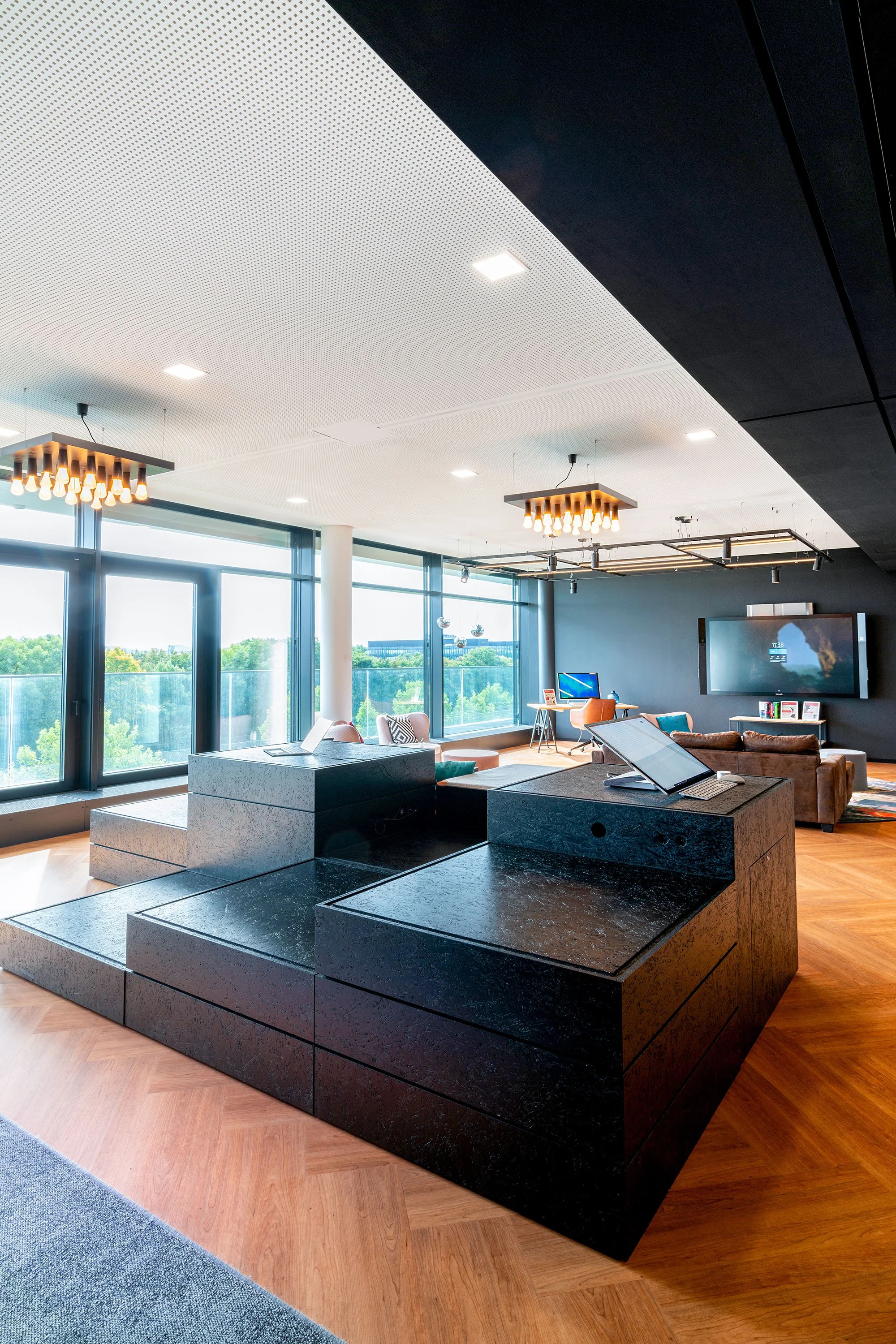 Modernes Büro mit schwarzen und weißen Deckenplatten, großen Fenstern, bequemen Sitzmöbeln und zwei eleganten Chandelieren mit E27 Glühbirnen