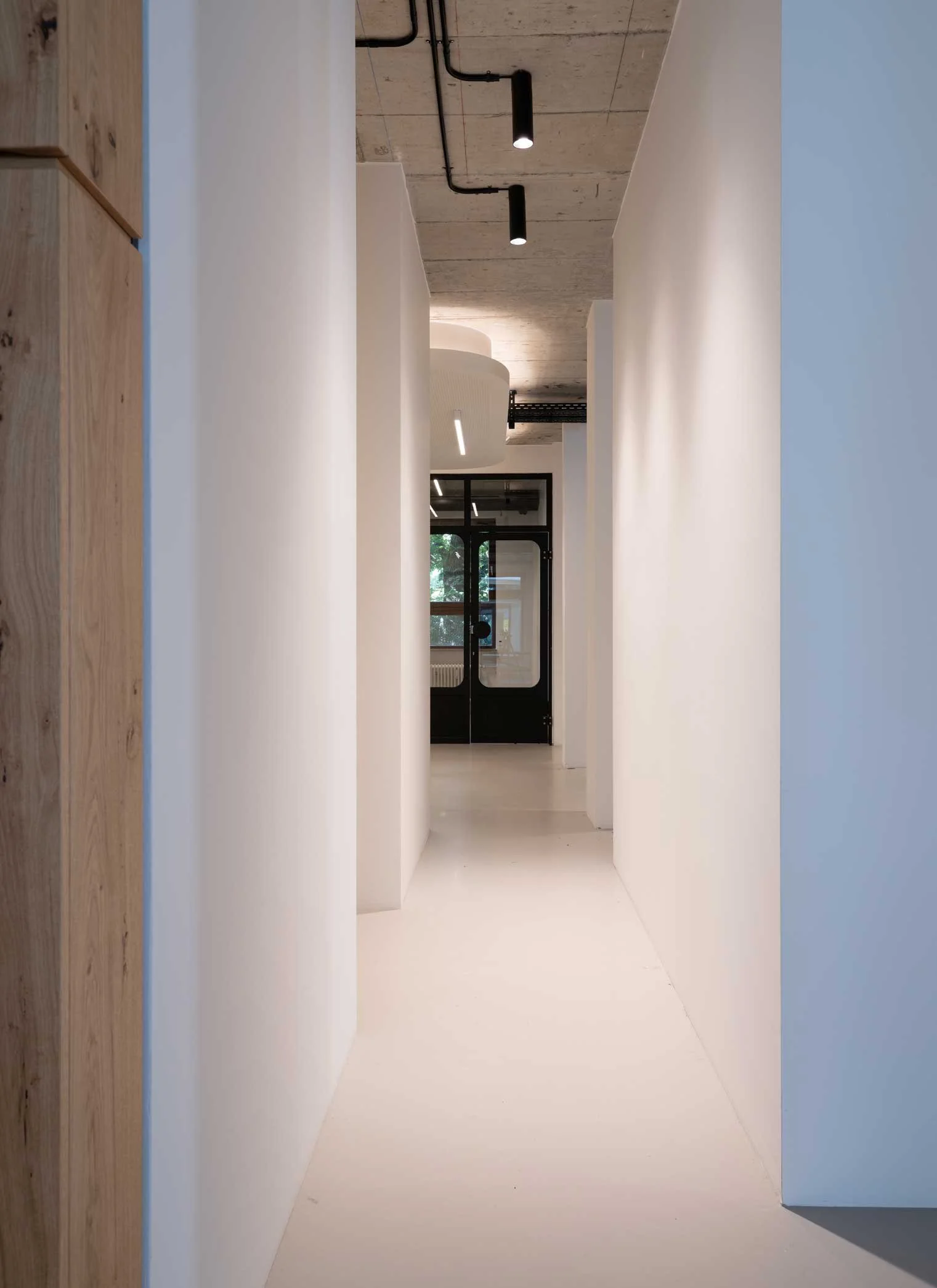 Ein moderner, minimalistisch gestalteter Flur mit weißen Wänden und schwarzer Deckenbeleuchtung in einem Gebäude mit großen Fenstern im Hintergrund.